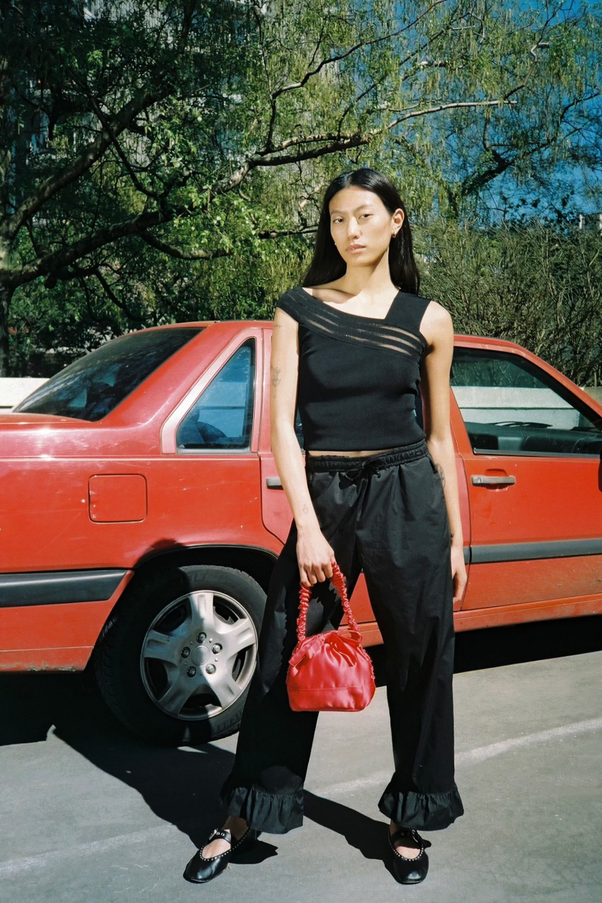 Woman in black outfit holding a red handbag standing next to a red car.