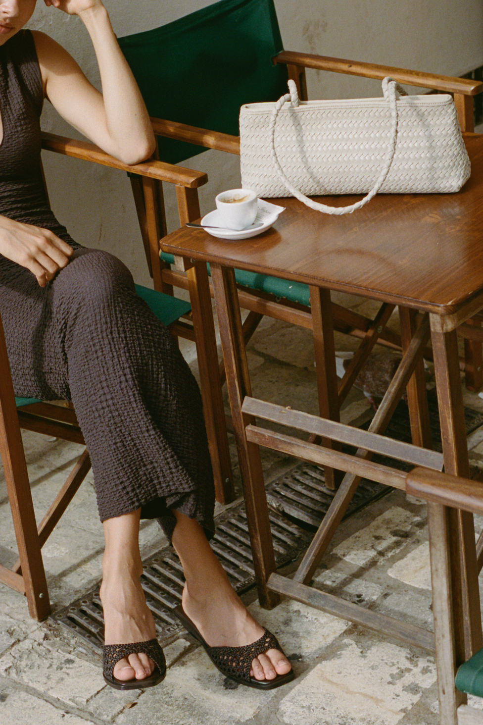 Woman sitting at a wooden table with a white handbag next to her, wearing brown shoes.