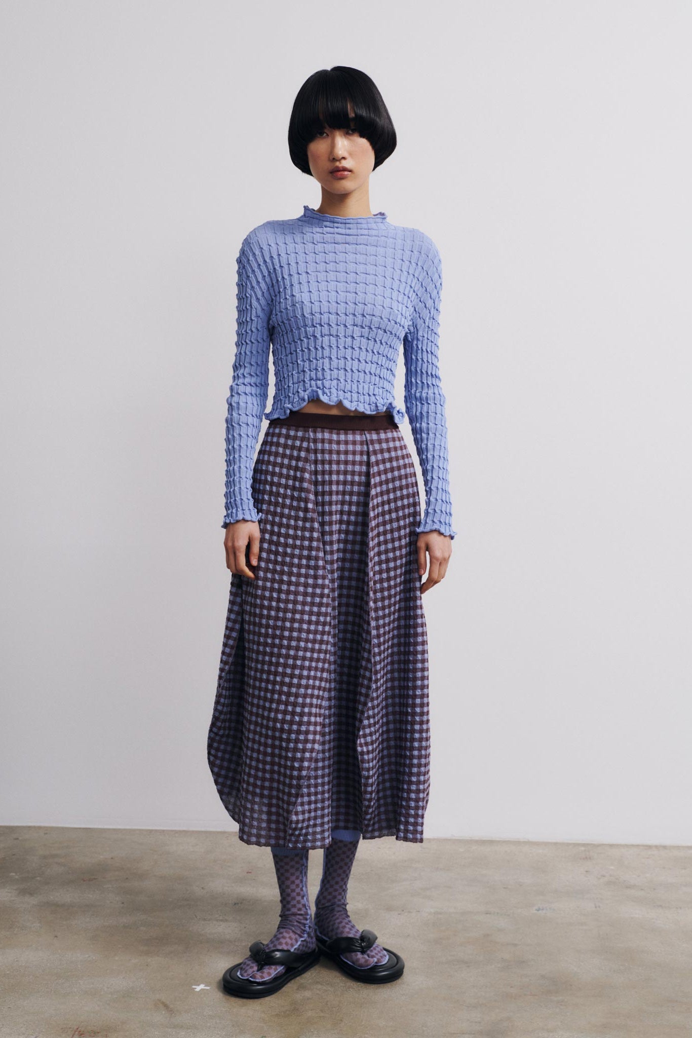 Person wearing a blue textured top and plaid skirt against a plain background