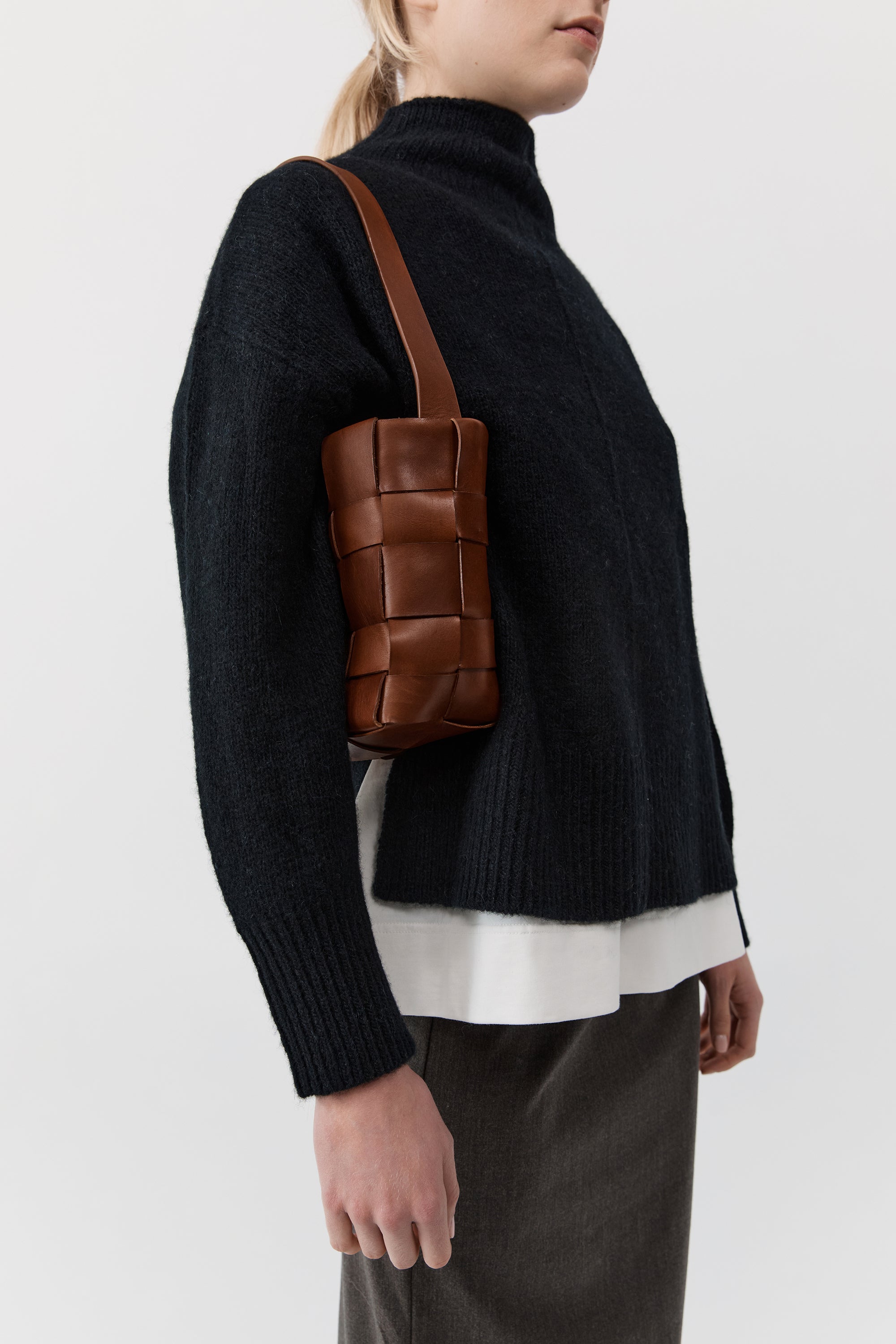 Person wearing a black sweater with a brown woven bag over one shoulder on a white background