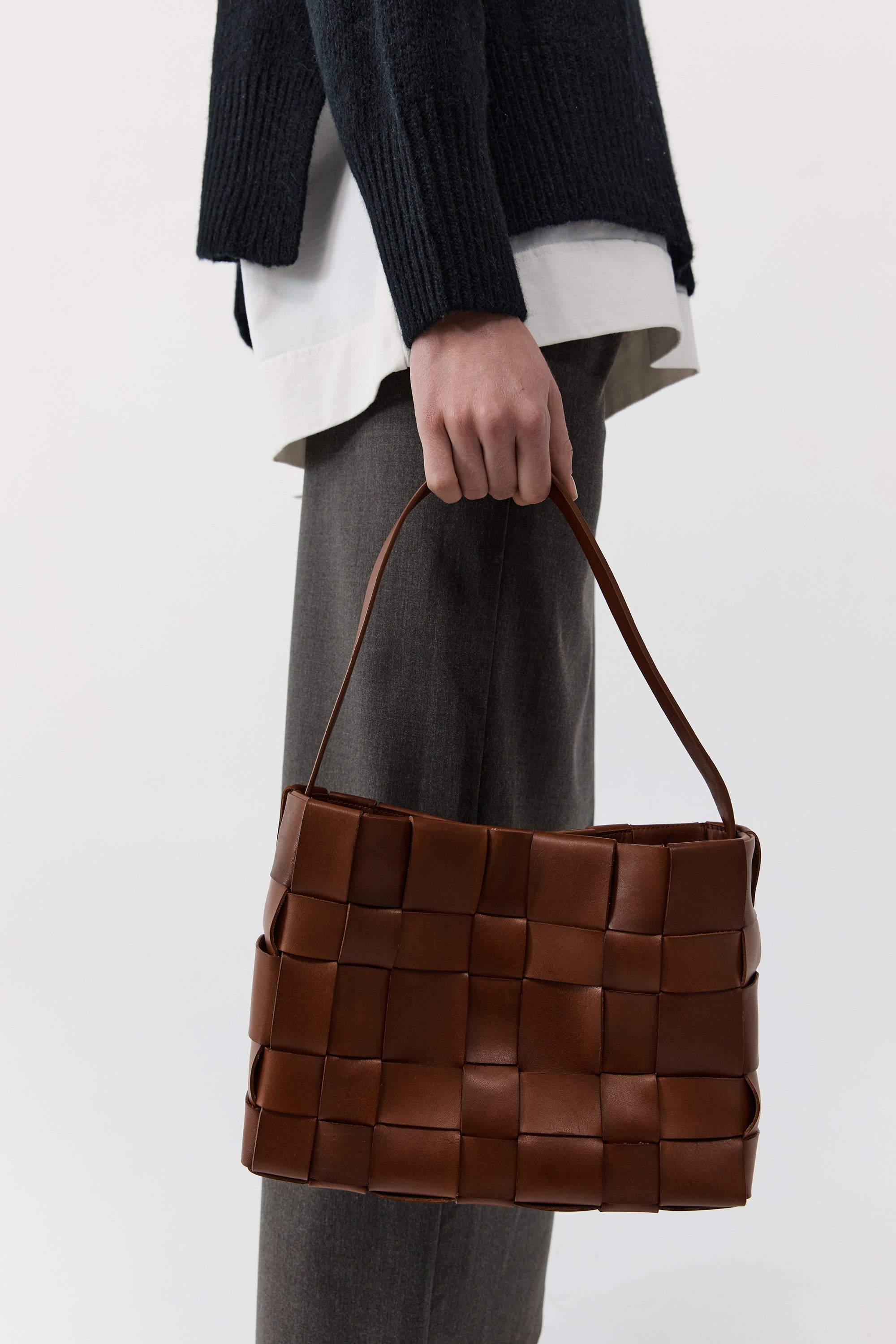 Brown woven handbag held by a person wearing a dark sweater and gray pants on a white background