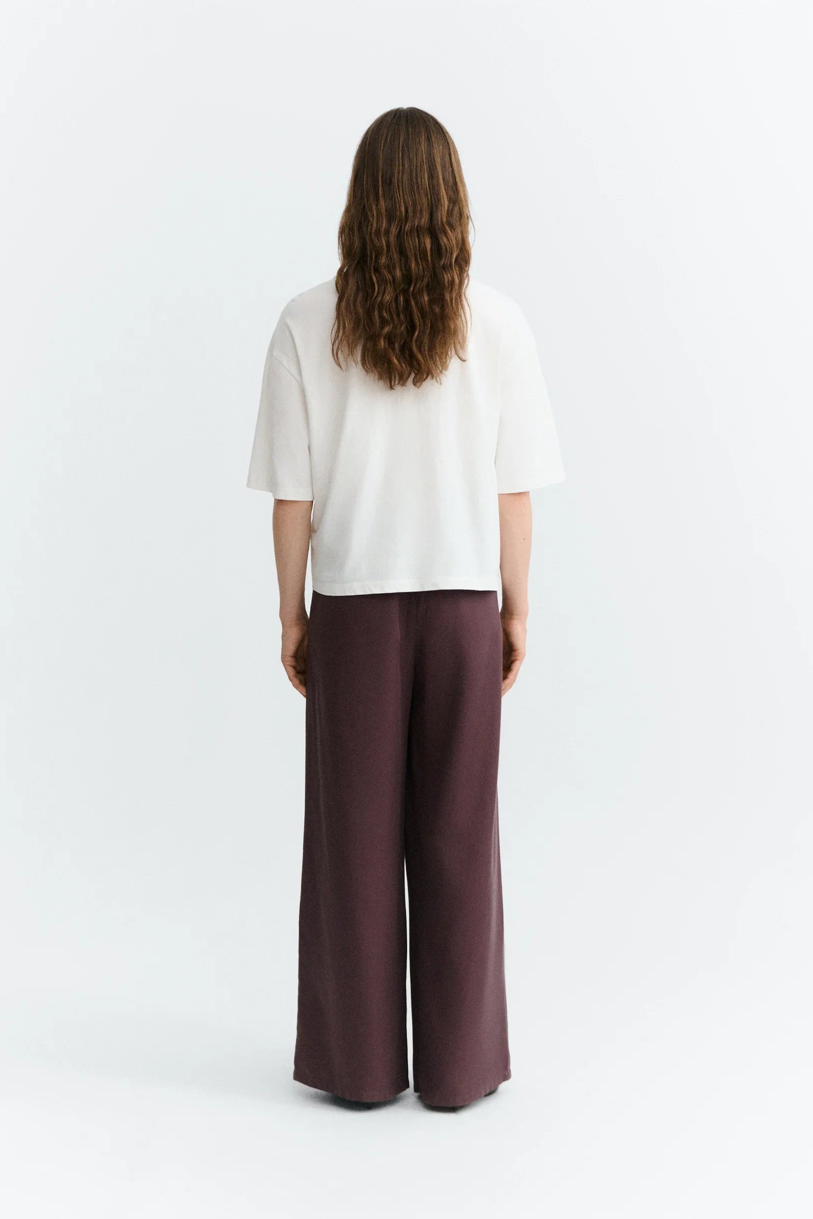 A woman viewed from behind wearing a white short-sleeve top and dark plum wide-leg trousers, standing against a plain light gray studio background.