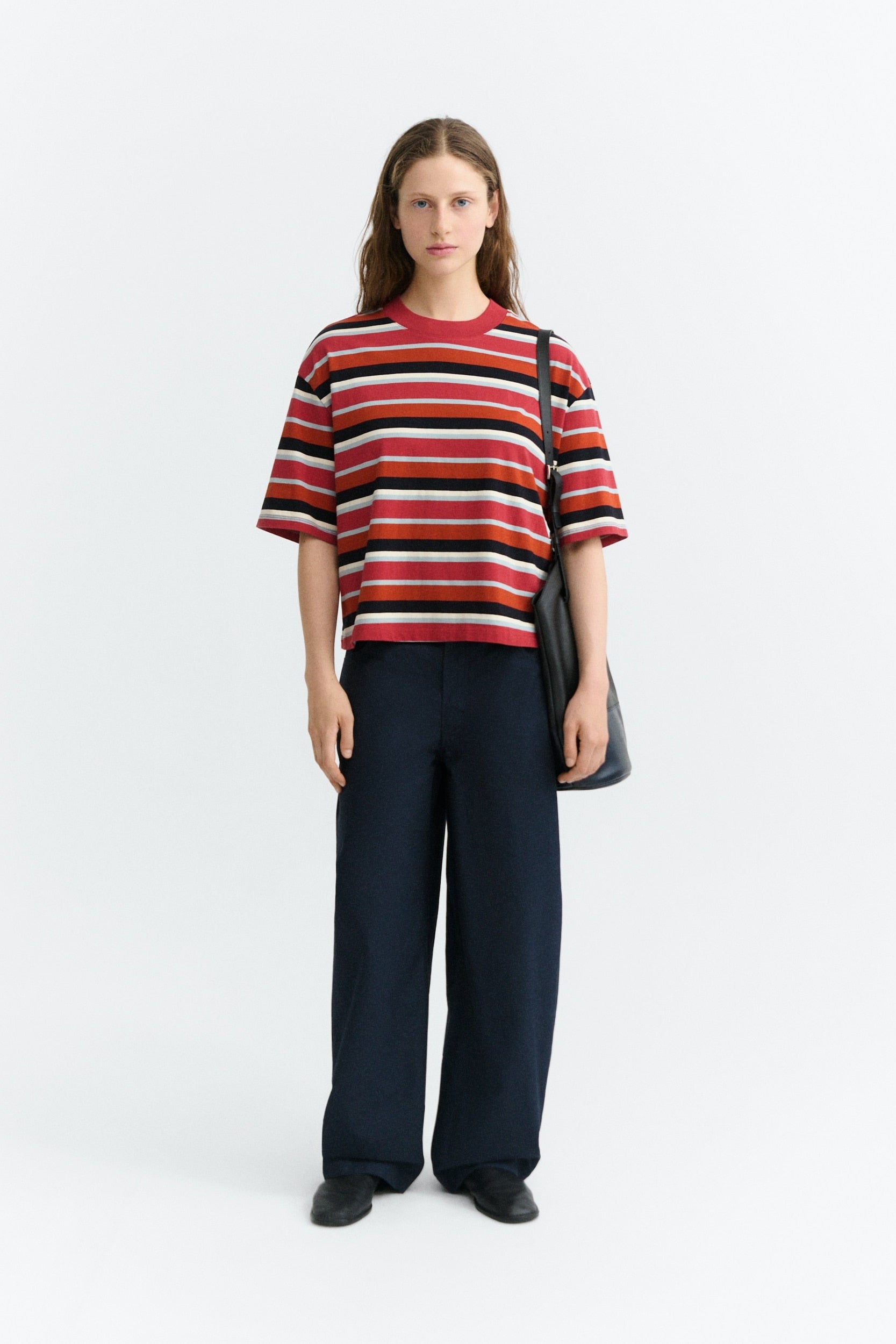 A model stands against a light gray studio background wearing a short-sleeve knit striped t-shirt with red, black, white, and orange horizontal stripes, paired with dark navy wide-leg trousers and a black shoulder bag.