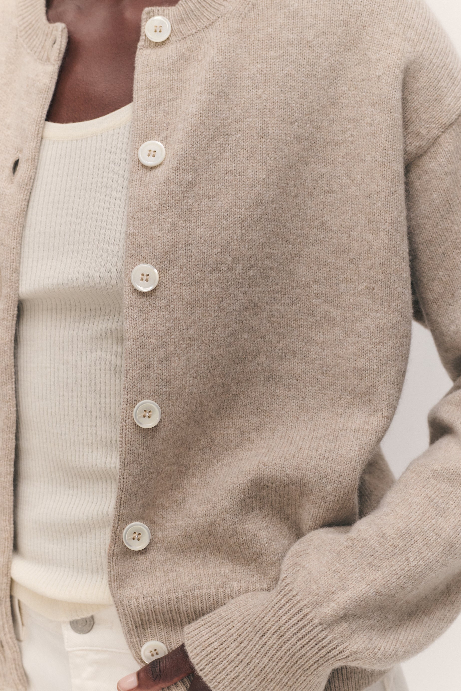 A close-up of a person wearing a light taupe knit cardigan over a cream ribbed top, with the cardigan featuring large cream buttons and ribbed cuffs and hem.