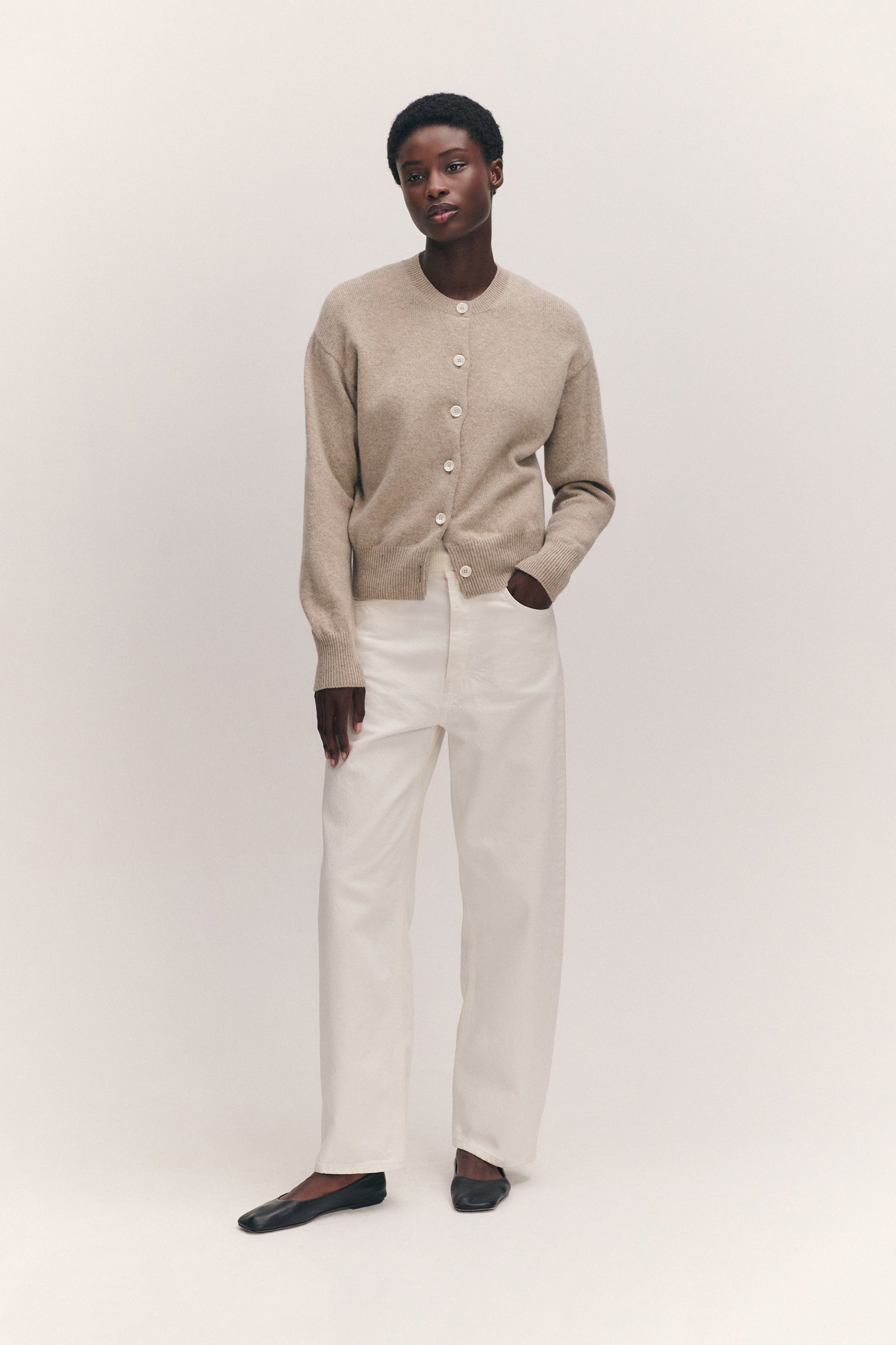 A model stands against a plain light beige studio background wearing a beige knit cardigan with white buttons, off-white wide-leg trousers, and dark flat shoes.