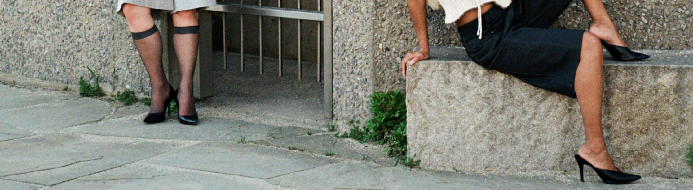 Two people in high heels on a sidewalk, one sitting on a stone bench.