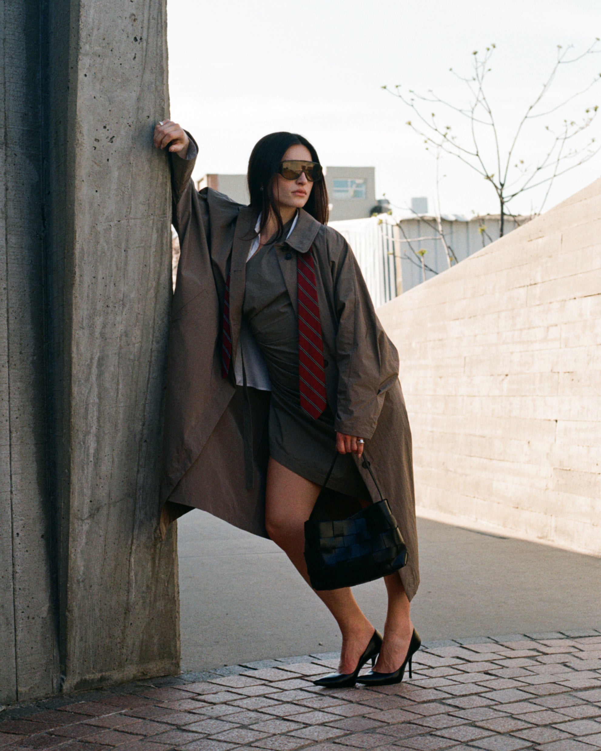 Woman in a long coat and sunglasses leaning against a wall outdoors.