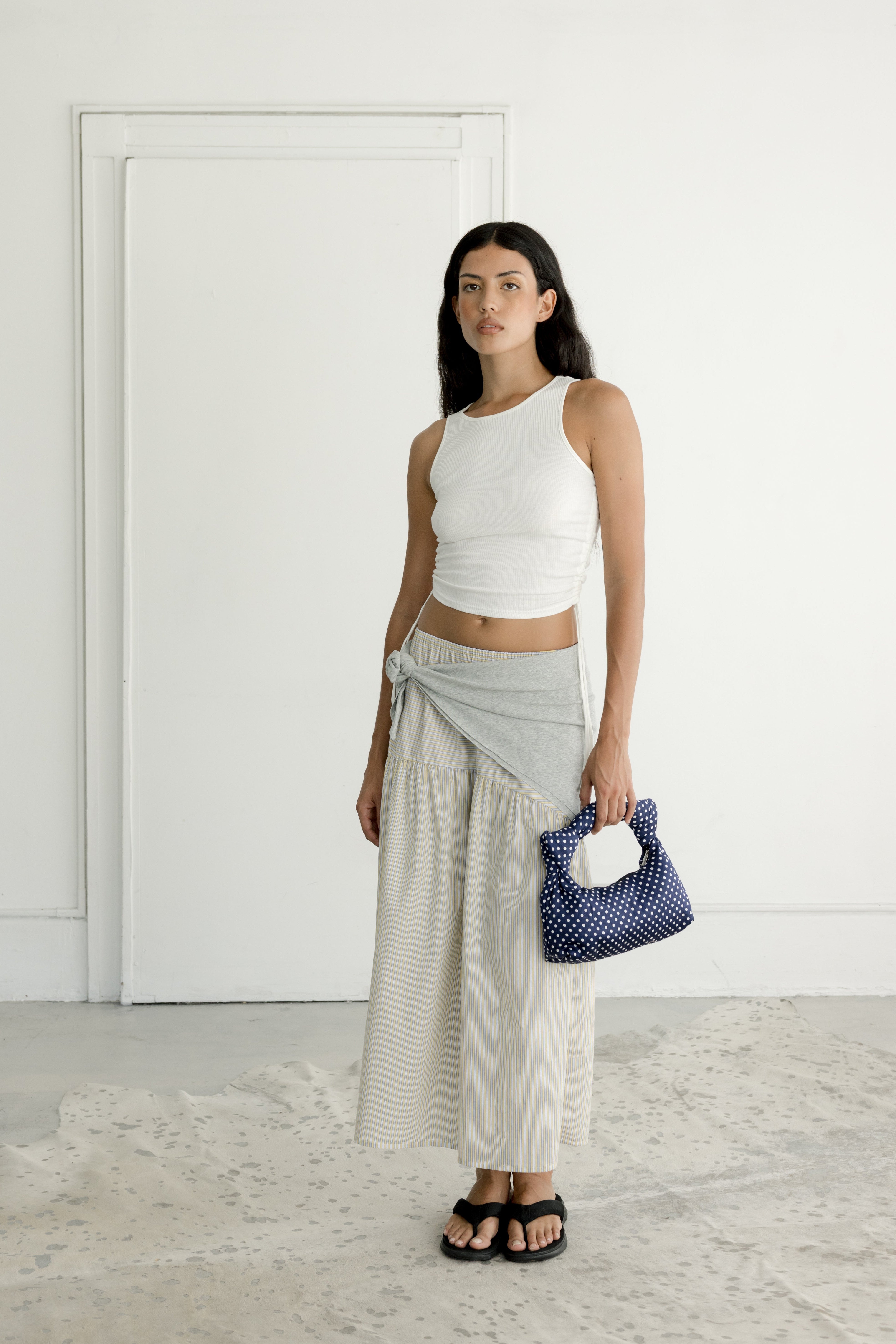 Woman wearing a white top and light gray skirt holding a blue handbag in a minimalistic setting.