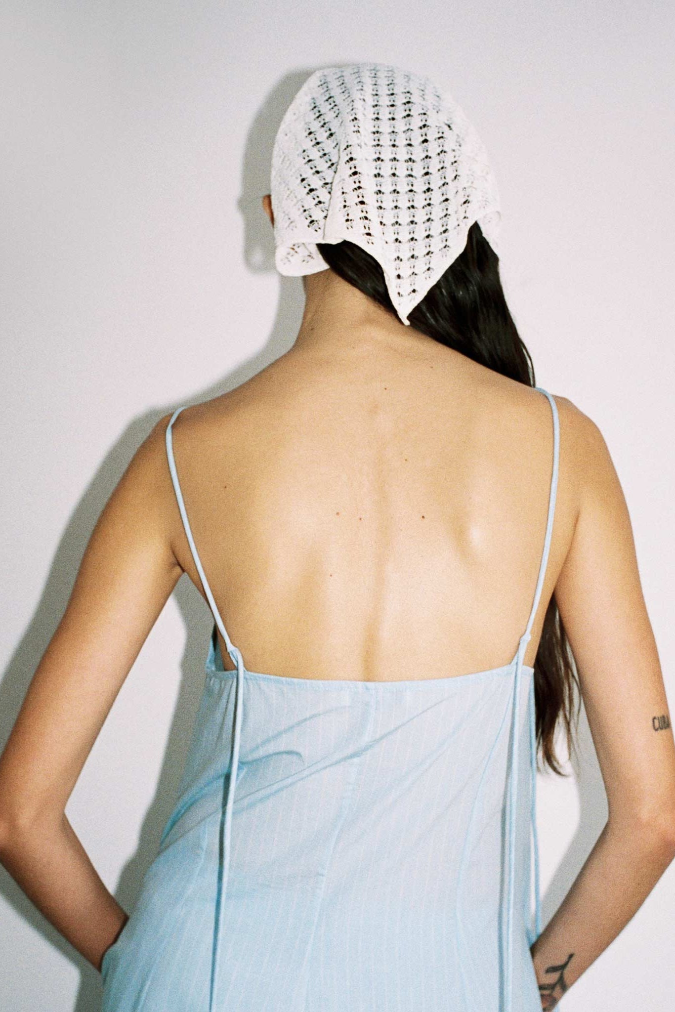 Person wearing a light blue dress with thin straps and a white headscarf, standing against a plain background.