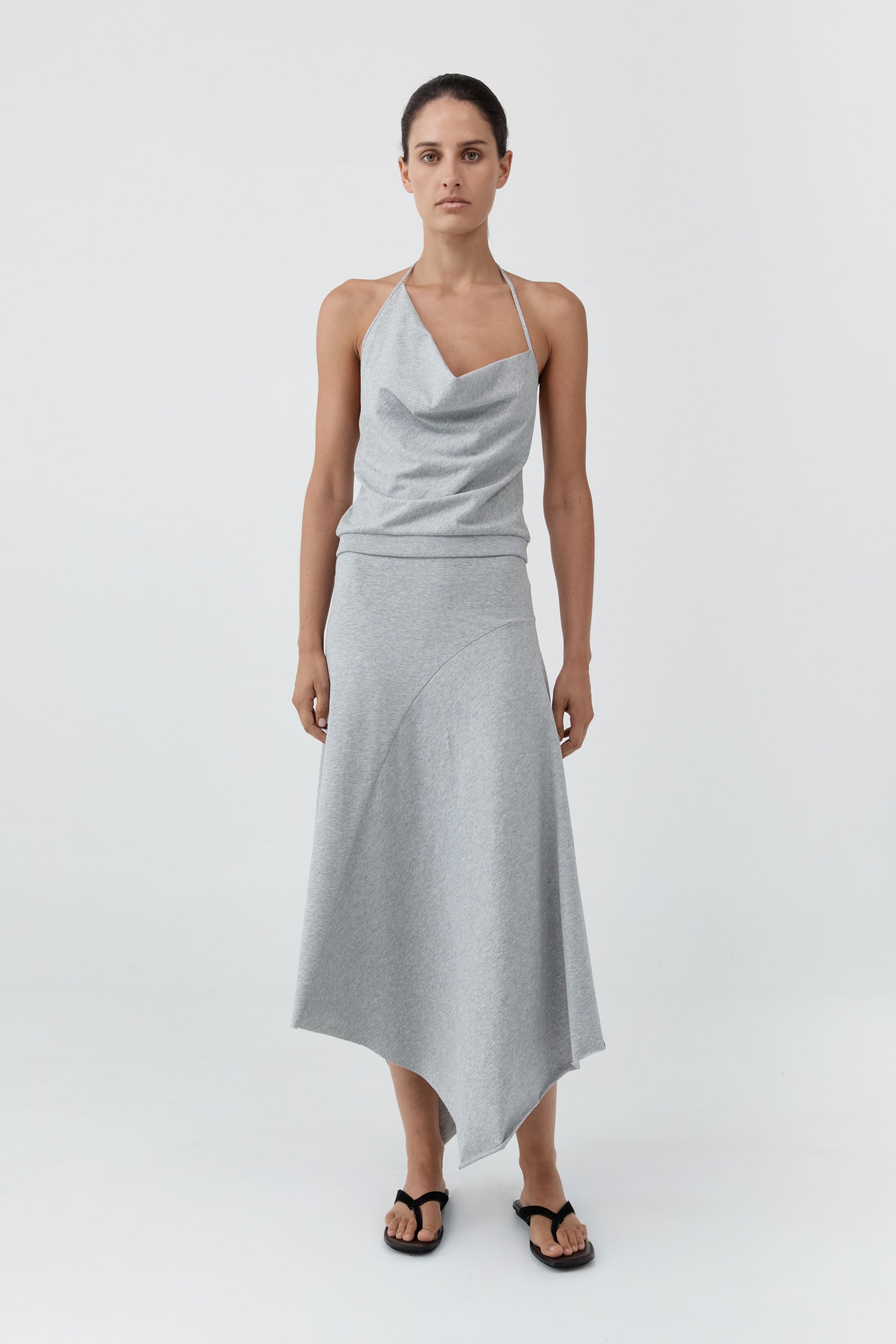 A model wearing a light gray asymmetrical draped skirt with a halter top with a draped cowl neckline and a high-low hemline, paired with black sandals, against a plain light gray studio background.