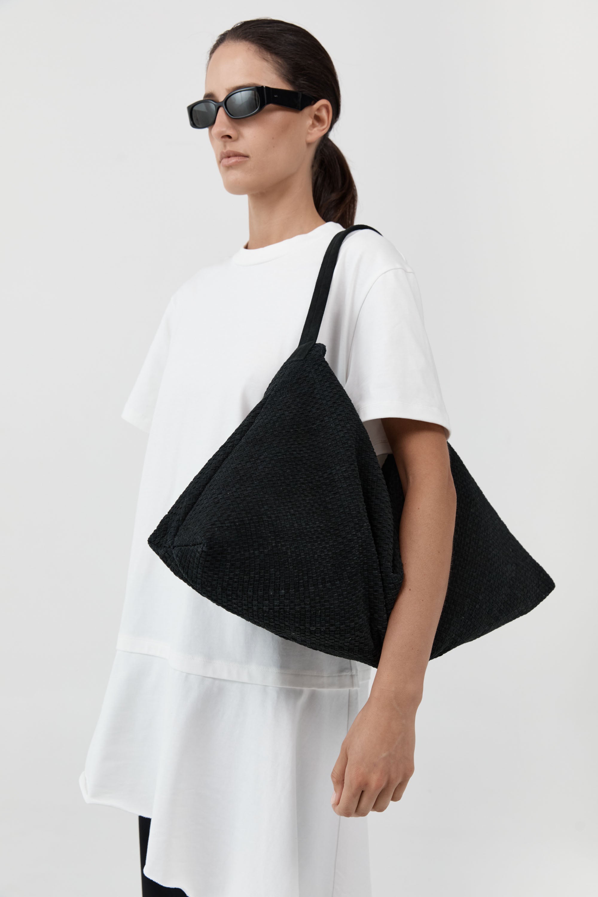 A model wearing a white oversized t-shirt and black sunglasses carries a large black woven shoulder bag against a light gray studio background.