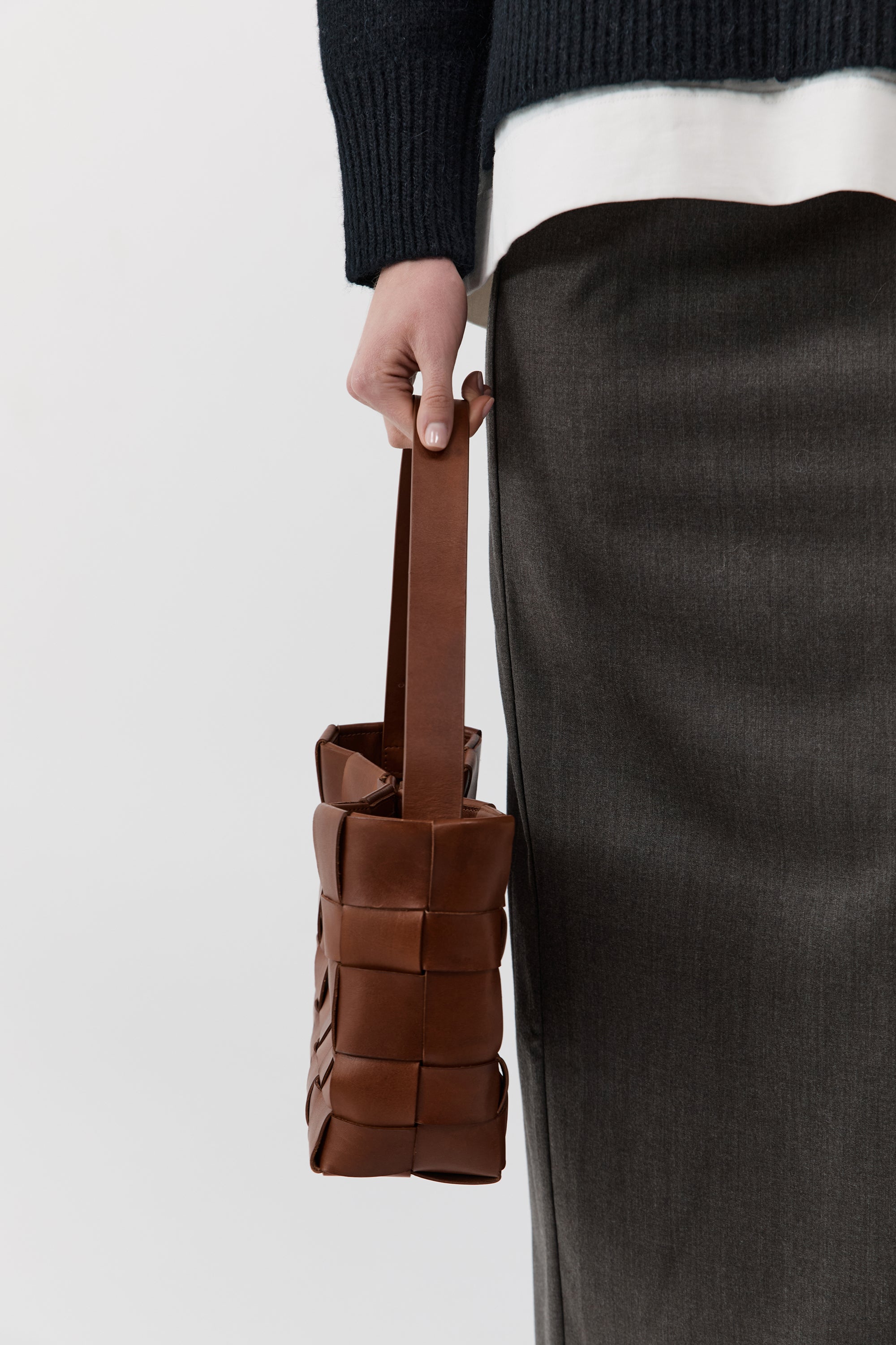 Brown woven handbag held by a person wearing a black sweater and gray pants on a white background
