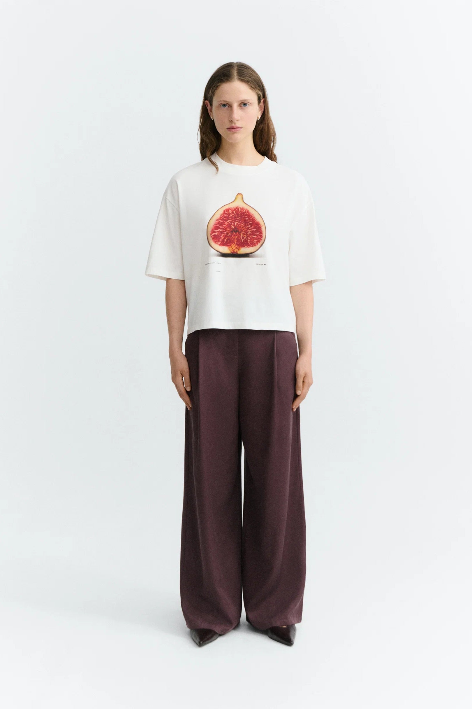 A model stands against a light gray studio background wearing a white cropped T-shirt with a large pink fig graphic on the chest and dark plum wide-leg pants.