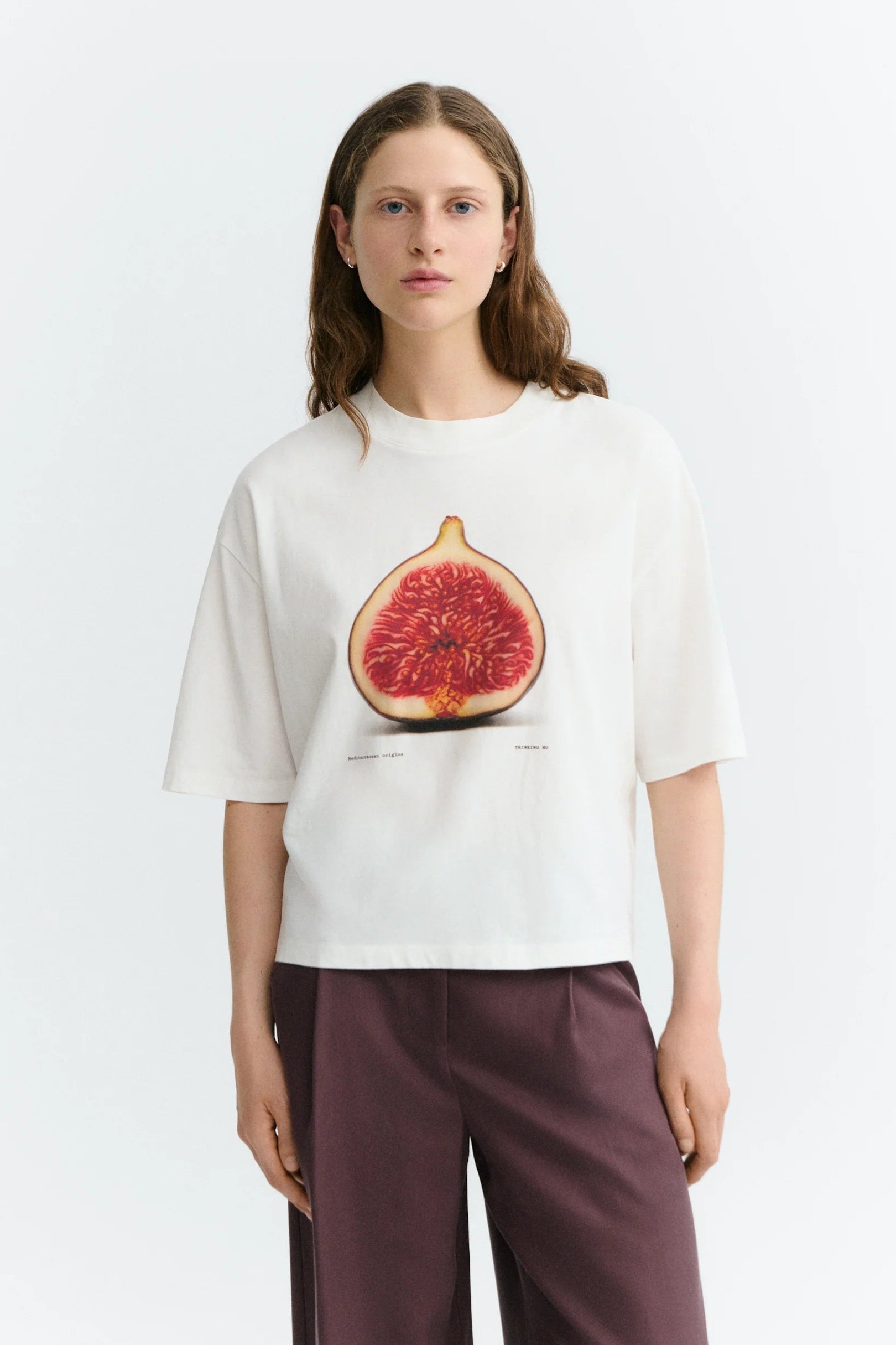 A woman modeling a white short-sleeve t-shirt with a large graphic print of a sliced fig on the front, paired with dark purple wide-leg pants, against a plain light gray background.