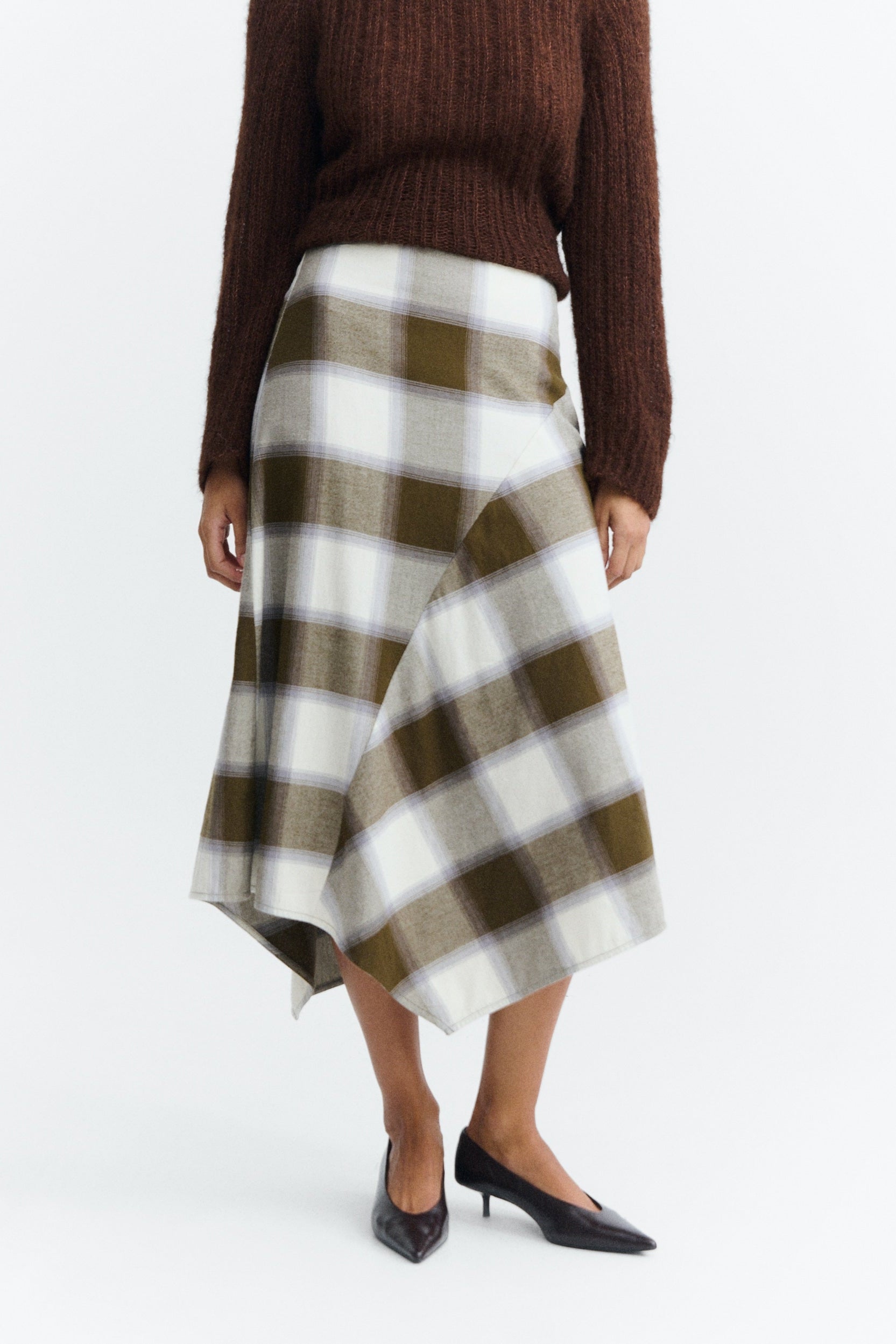 A model wearing a brown ribbed knit sweater and a high-waisted, asymmetrical plaid skirt in brown, cream, and white, paired with black pointed-toe heels.