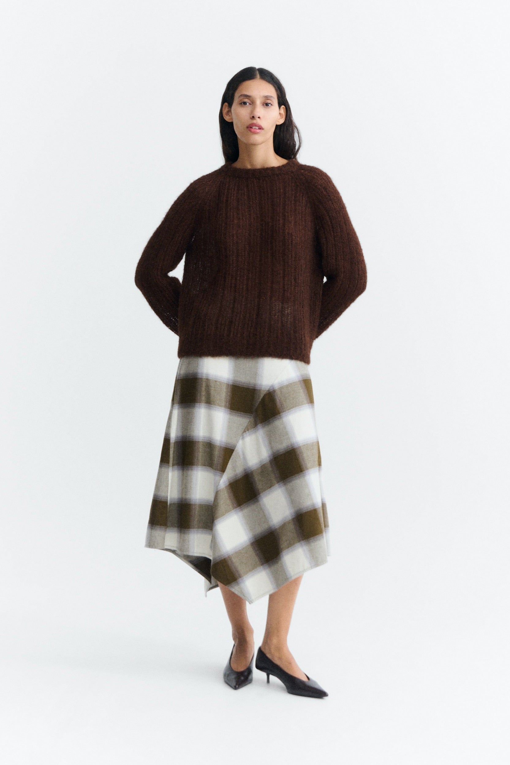 A model stands against a plain white studio background wearing a dark brown ribbed knit sweater, a brown, white, and gray plaid asymmetrical skirt, and black pointed-toe heels.