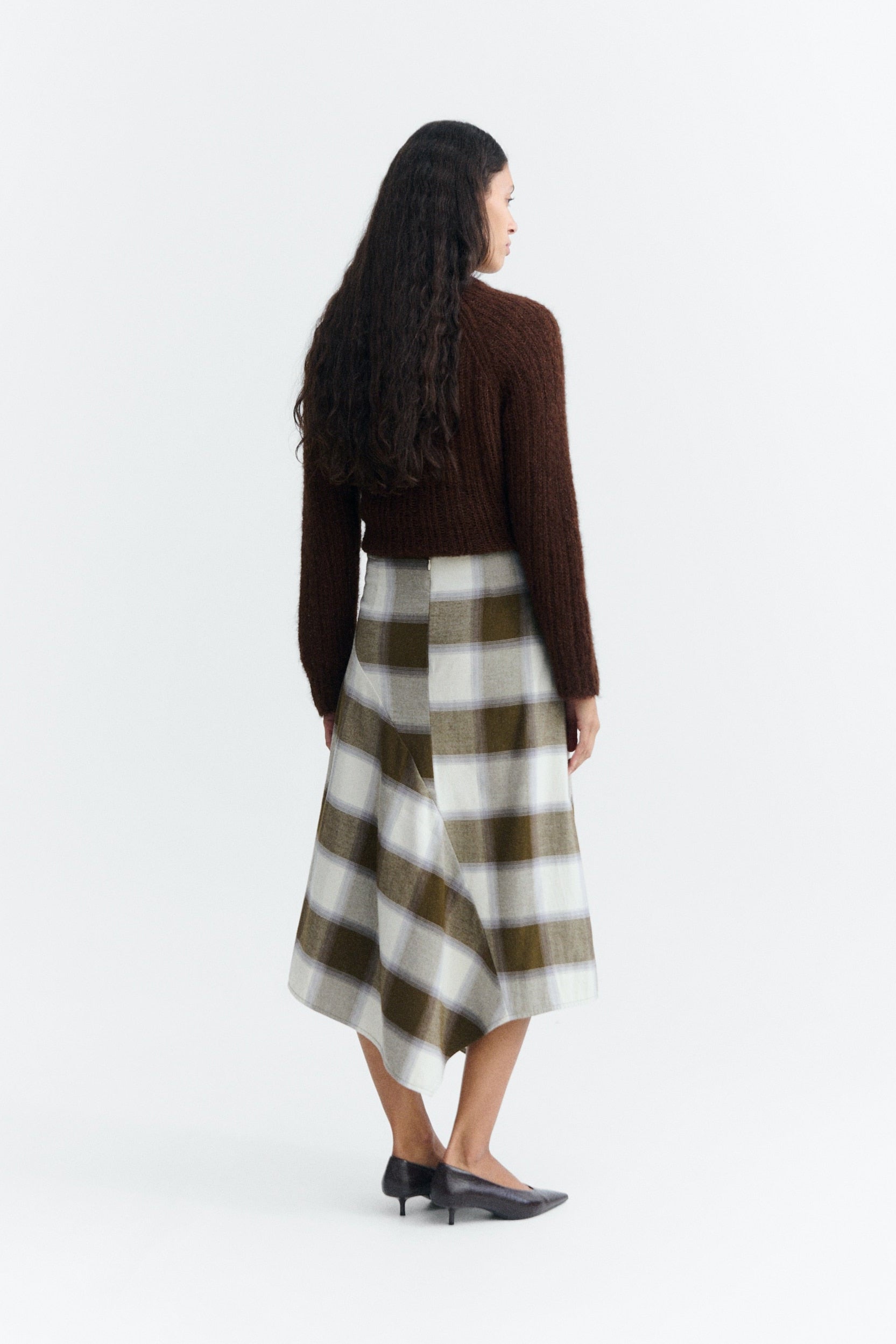 A woman is shown from the back wearing a dark brown knit sweater and a brown, white, and gray plaid midi skirt, paired with black pointed-toe flats, against a plain white studio background.