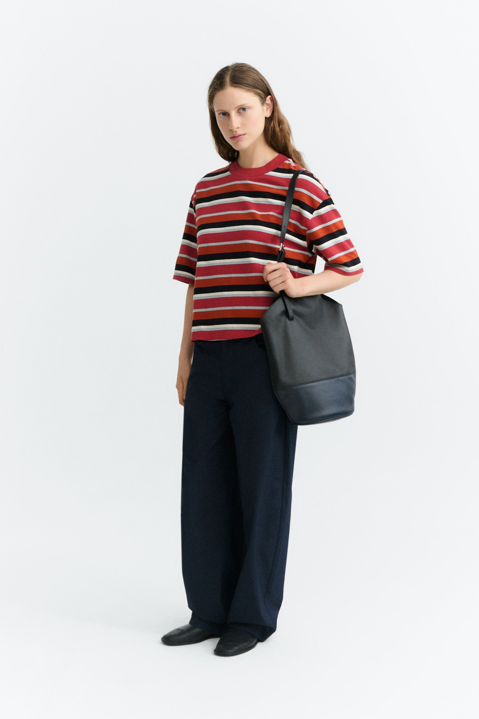 A woman stands against a plain light gray studio background wearing a short-sleeve striped knit top with red, navy, black, and white horizontal stripes, dark wide-leg trousers, and black shoes, while carrying a large dark gray shoulder bag.
