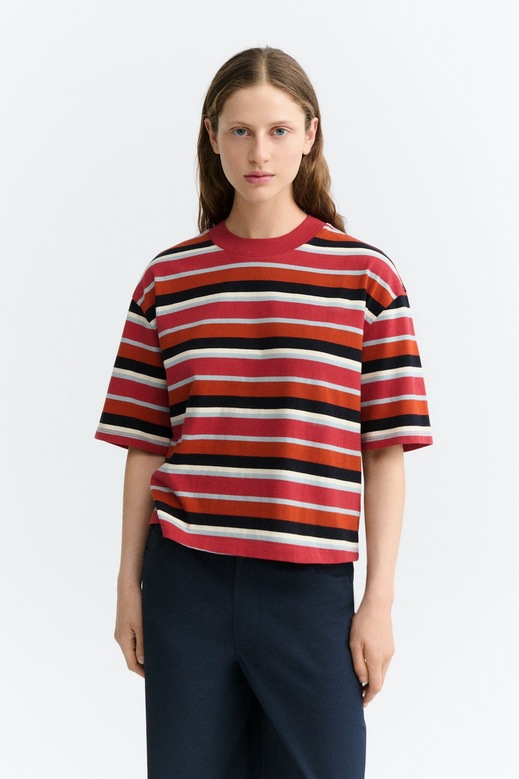 A woman modeling a short-sleeve crewneck knit top with horizontal stripes in red, orange, black, and off-white, paired with dark navy trousers, against a light gray studio background.