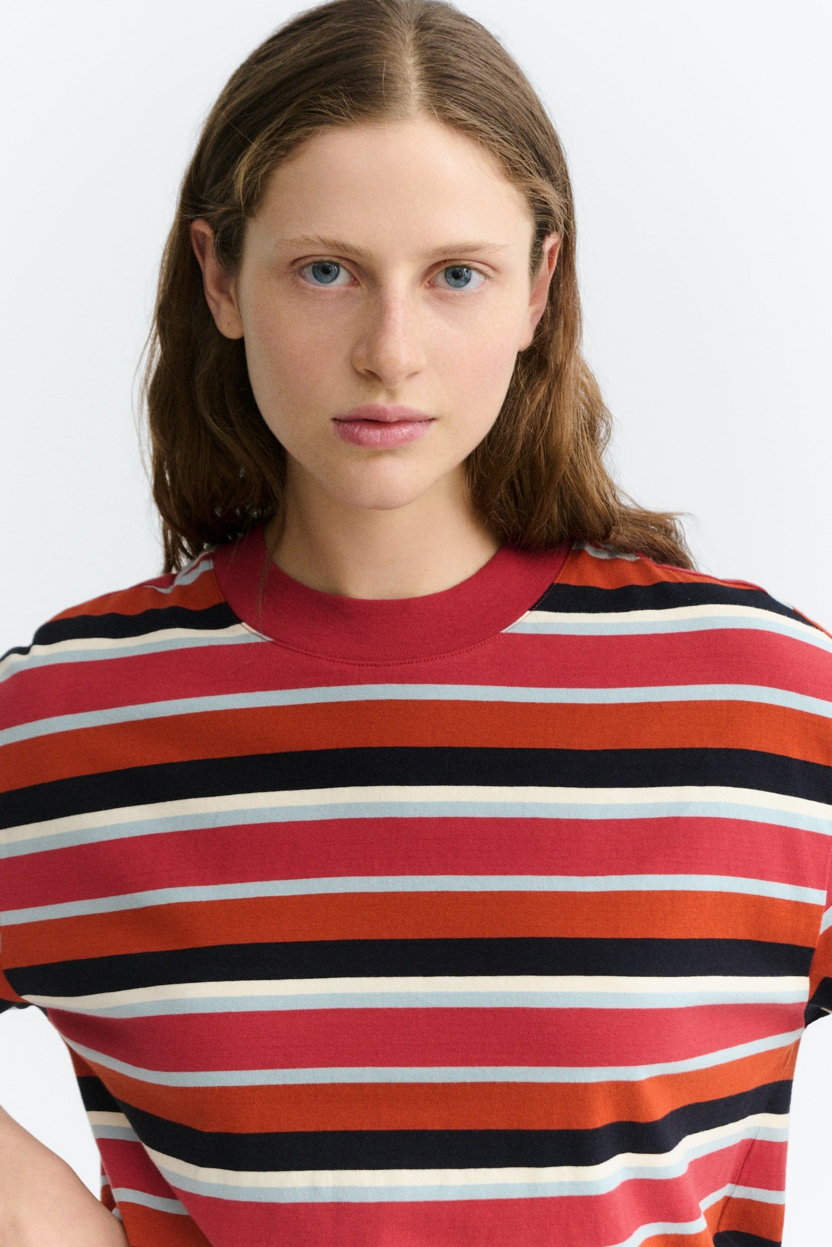 A woman wearing a short-sleeve crewneck t-shirt with horizontal stripes in red, orange, black, and white, photographed against a light gray studio background.