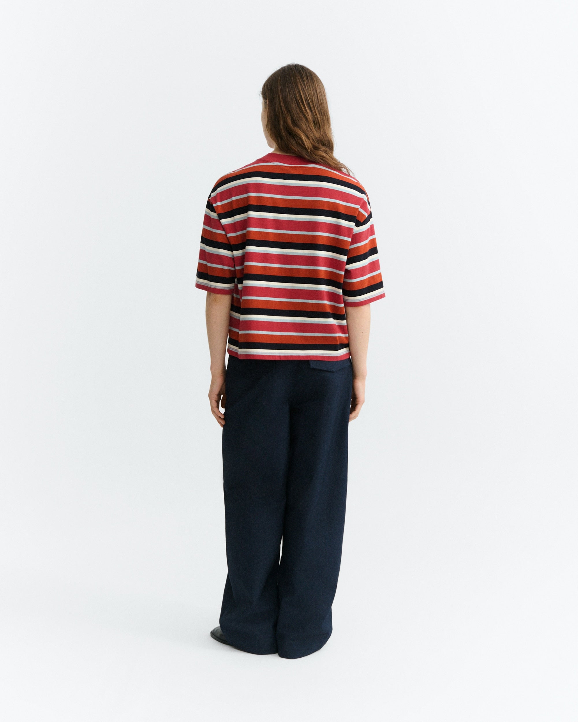 A model viewed from behind wearing a short-sleeve knit striped shirt with red, black, and white horizontal stripes, paired with dark navy wide-leg trousers, against a plain white studio background.