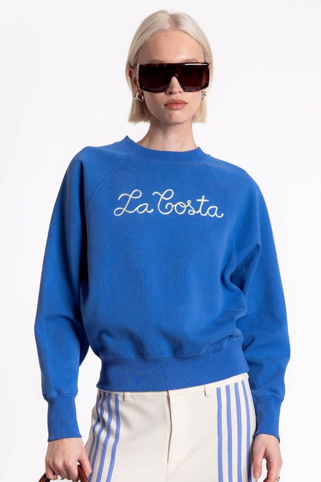 Woman wearing a blue sweatshirt with 'La Corte' text, white pants with blue stripes, and holding a brown handbag on a white background.