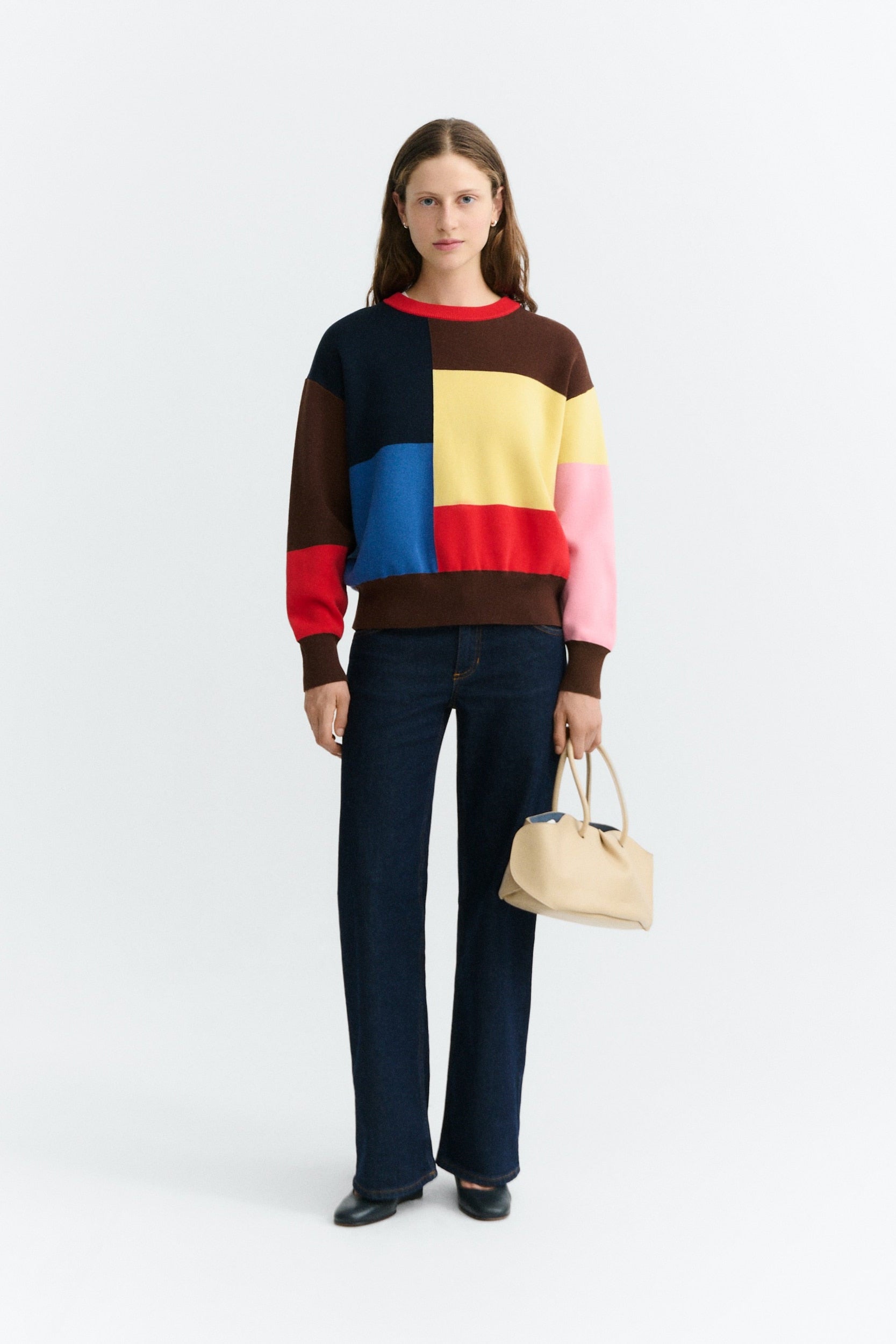 A model stands against a light gray studio background wearing a color-block knit sweater with dark blue jeans and holding a beige handbag.