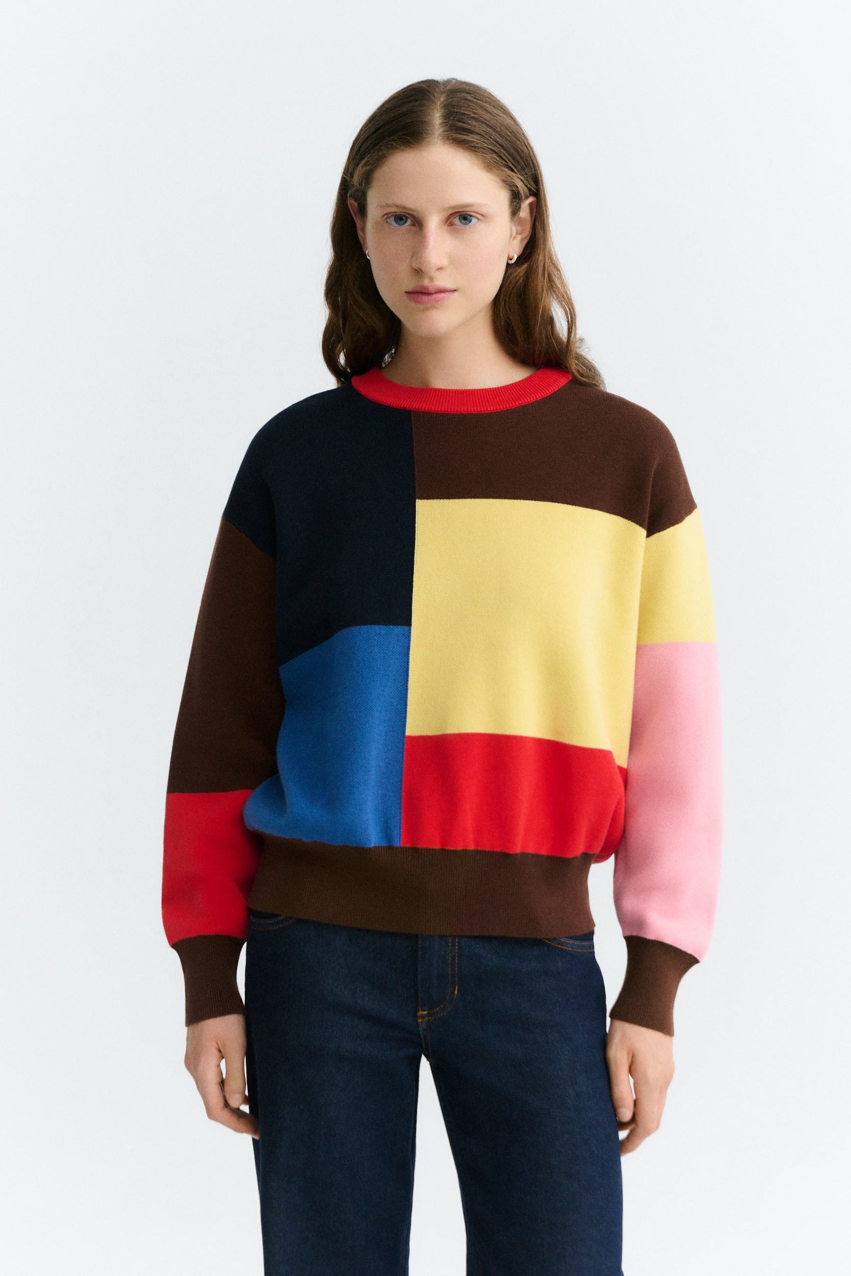 A woman modeling a color-block crewneck sweatshirt with blocks of navy, brown, yellow, pink, and red, paired with dark blue jeans against a light gray background.
