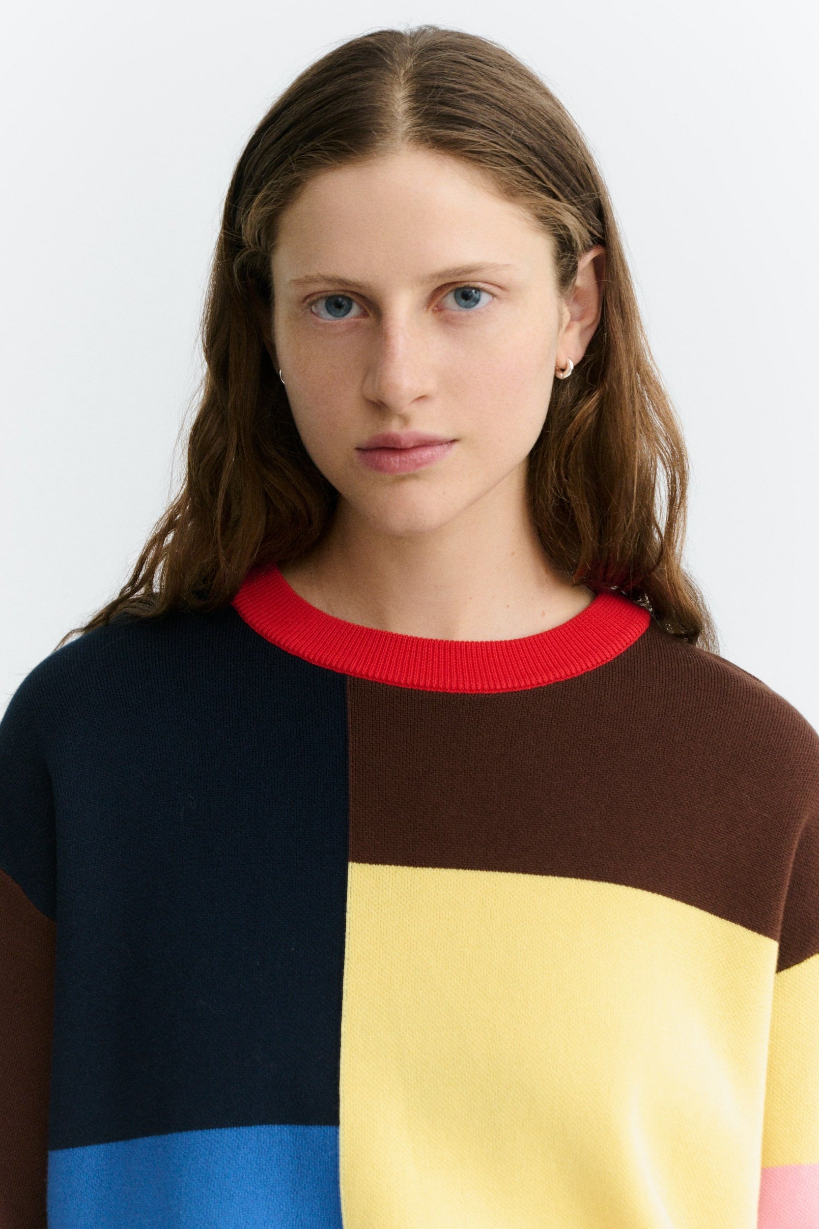 A woman wearing a color-block knit sweater with a red crew neck, navy blue, brown, yellow, and blue blocks, photographed against a light gray studio background.