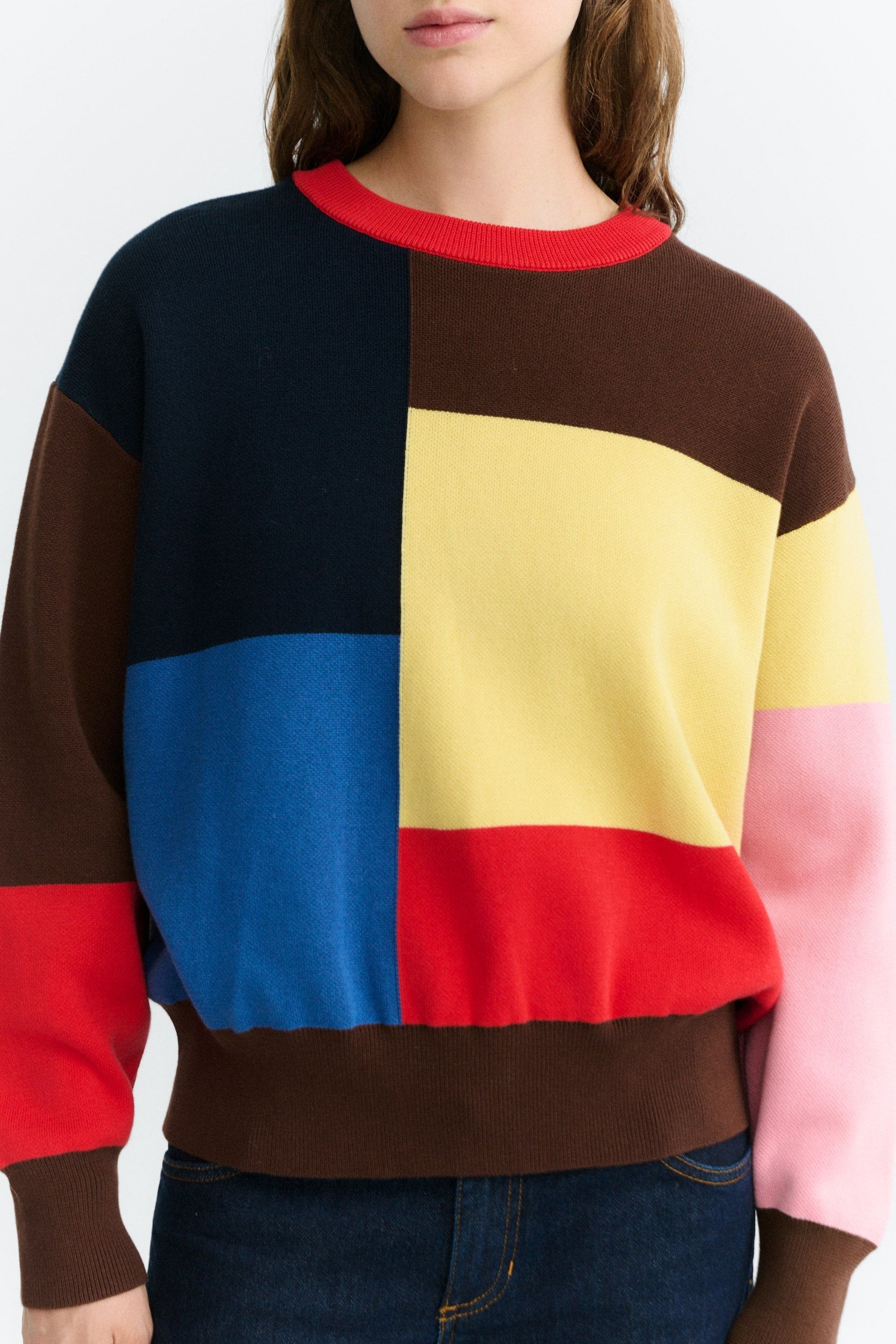 A woman wearing a color-block knit crewneck sweater with panels of navy, brown, yellow, blue, red, and pink, paired with dark blue jeans against a light gray background.