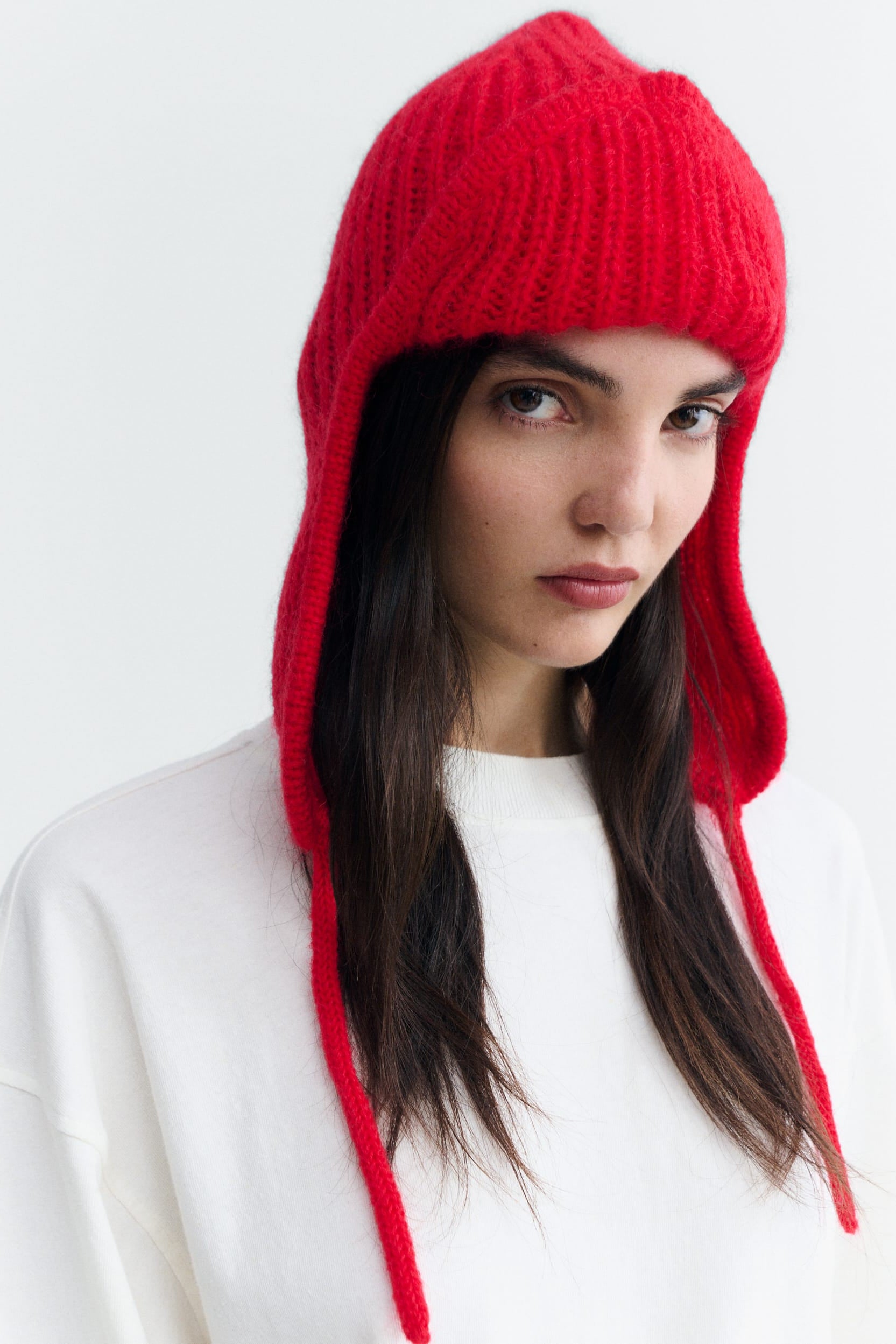A woman wearing a bright red knit hooded beanie with ear flaps and long ties, paired with a white sweatshirt, against a plain light gray background.