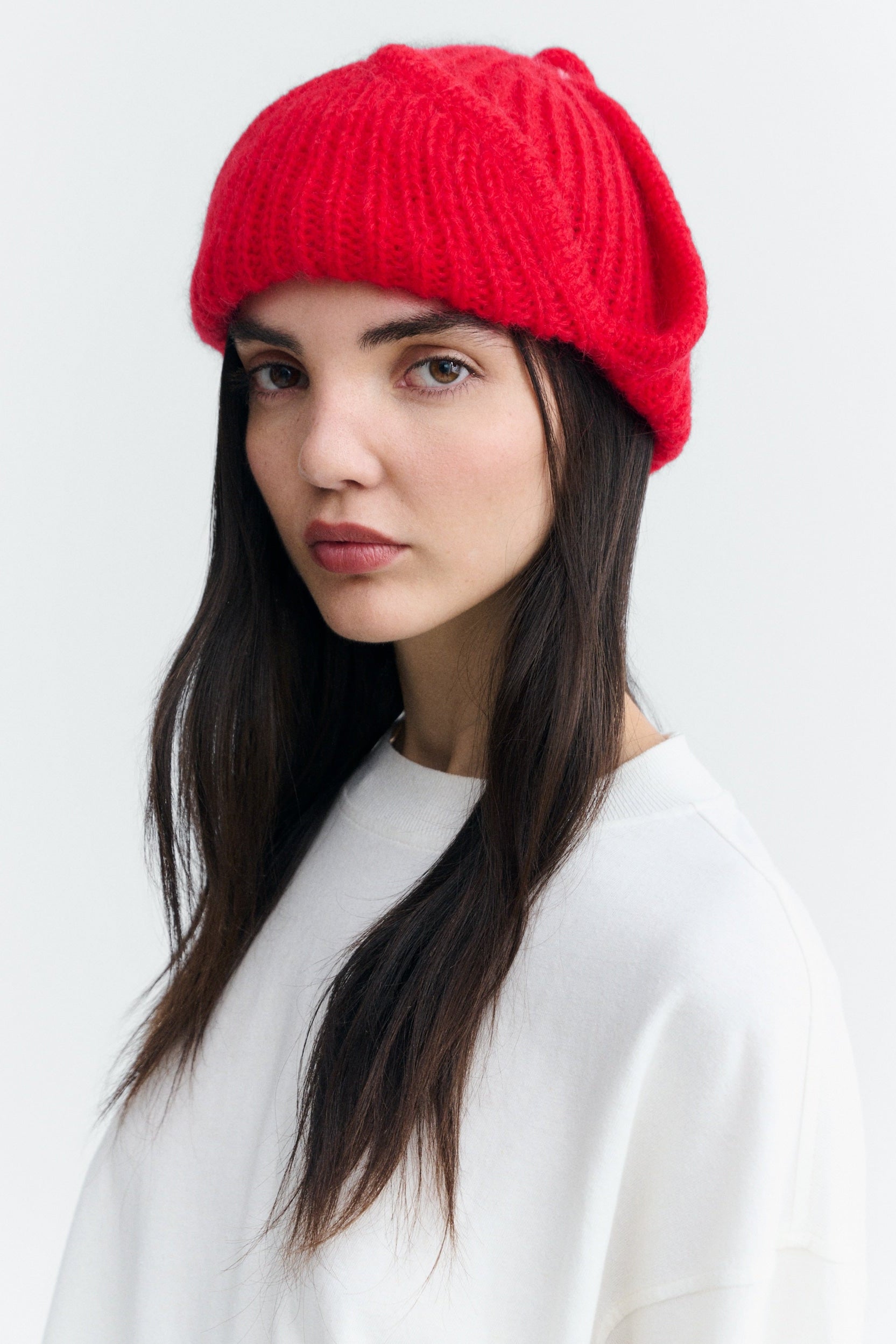 A woman with long dark hair wearing a bright red ribbed knit slouchy beanie and a white sweatshirt, photographed against a clean light gray studio background.