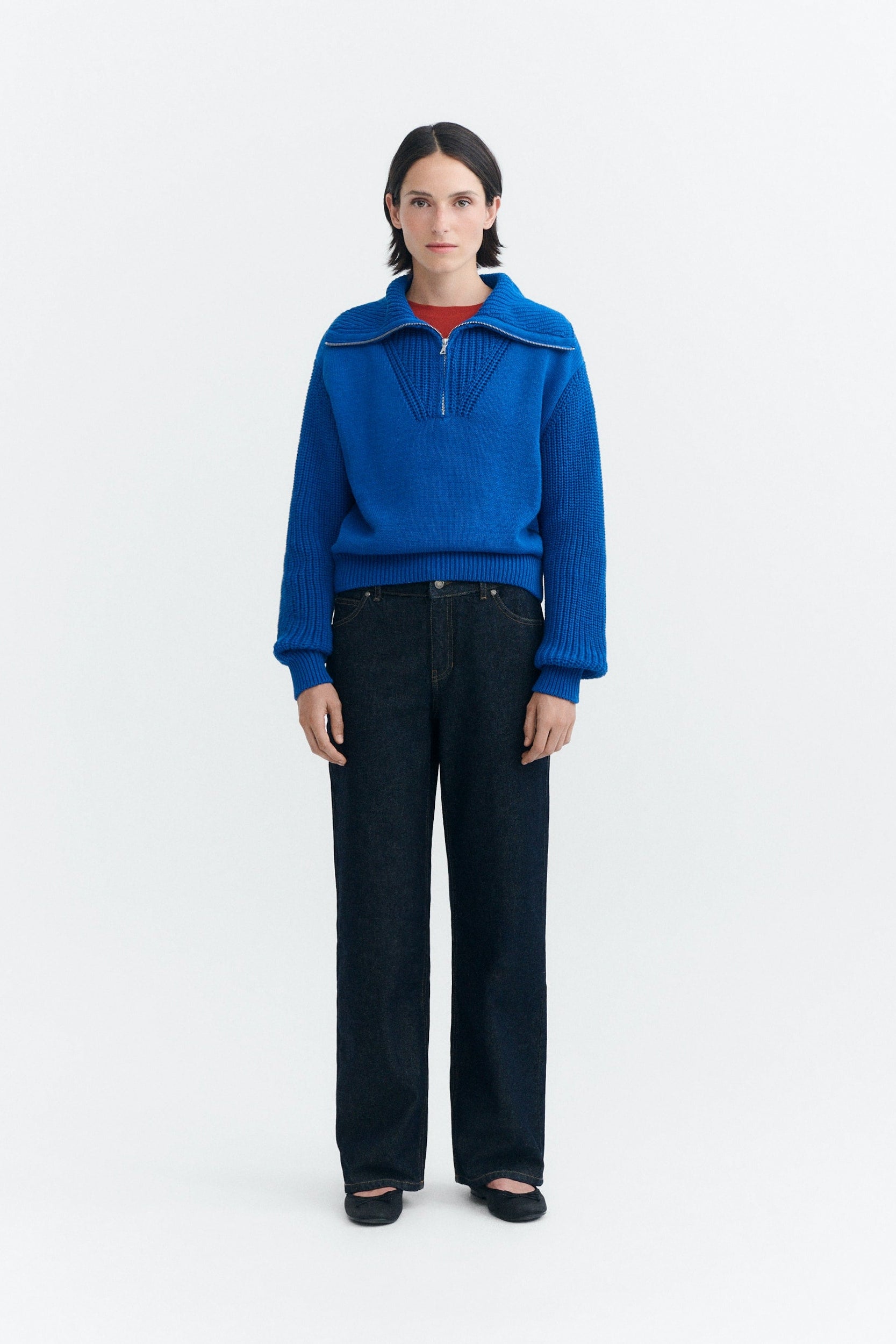 A model stands against a plain white studio background wearing a bright blue knit half-zip sweater with a wide collar, dark wide-leg jeans, and black flats.