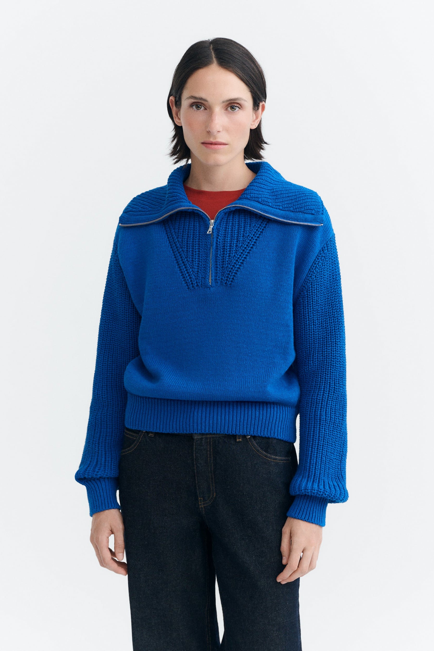A model wearing a vibrant blue ribbed knit half-zip sweater with a unique collar design, layered over a red top and dark jeans, against a plain white studio background.