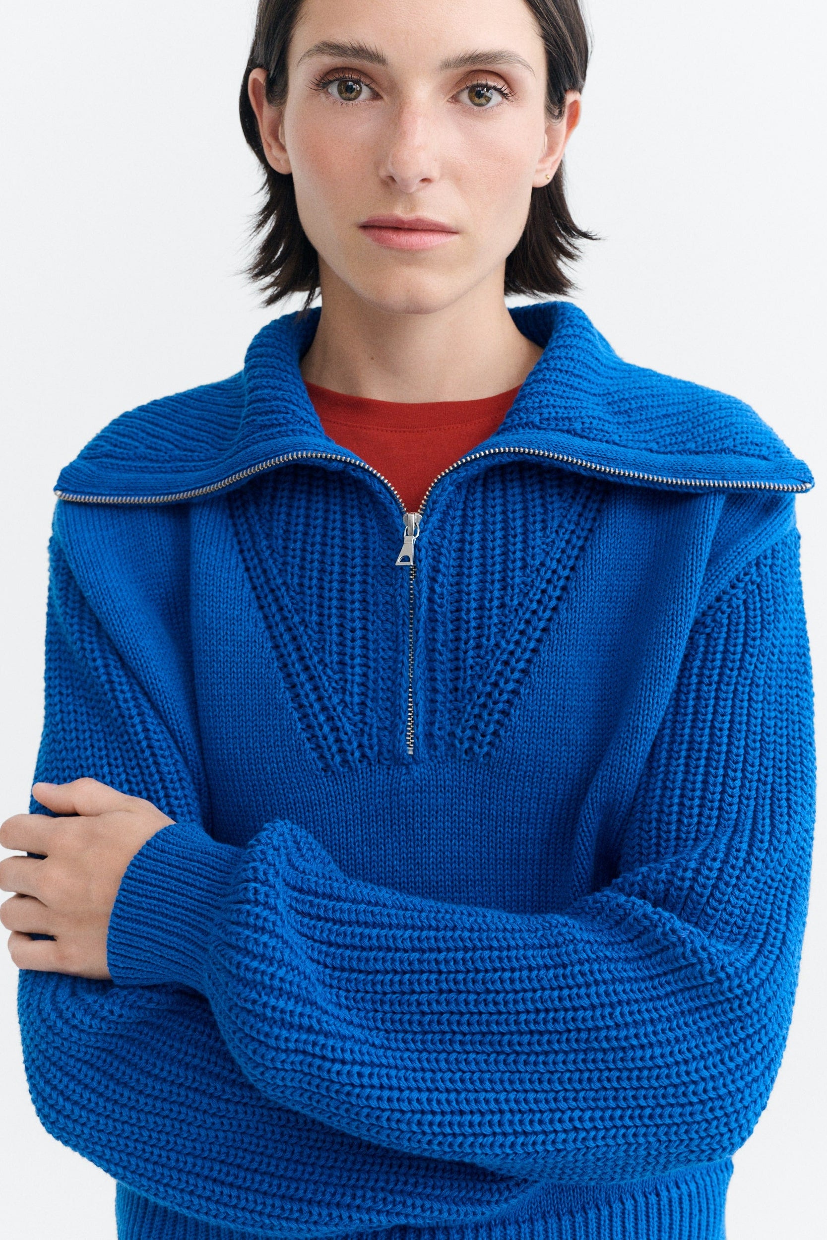 A model wearing a bright blue ribbed knit half-zip sweater with a wide collar, layered over a red crewneck shirt, against a plain white background.