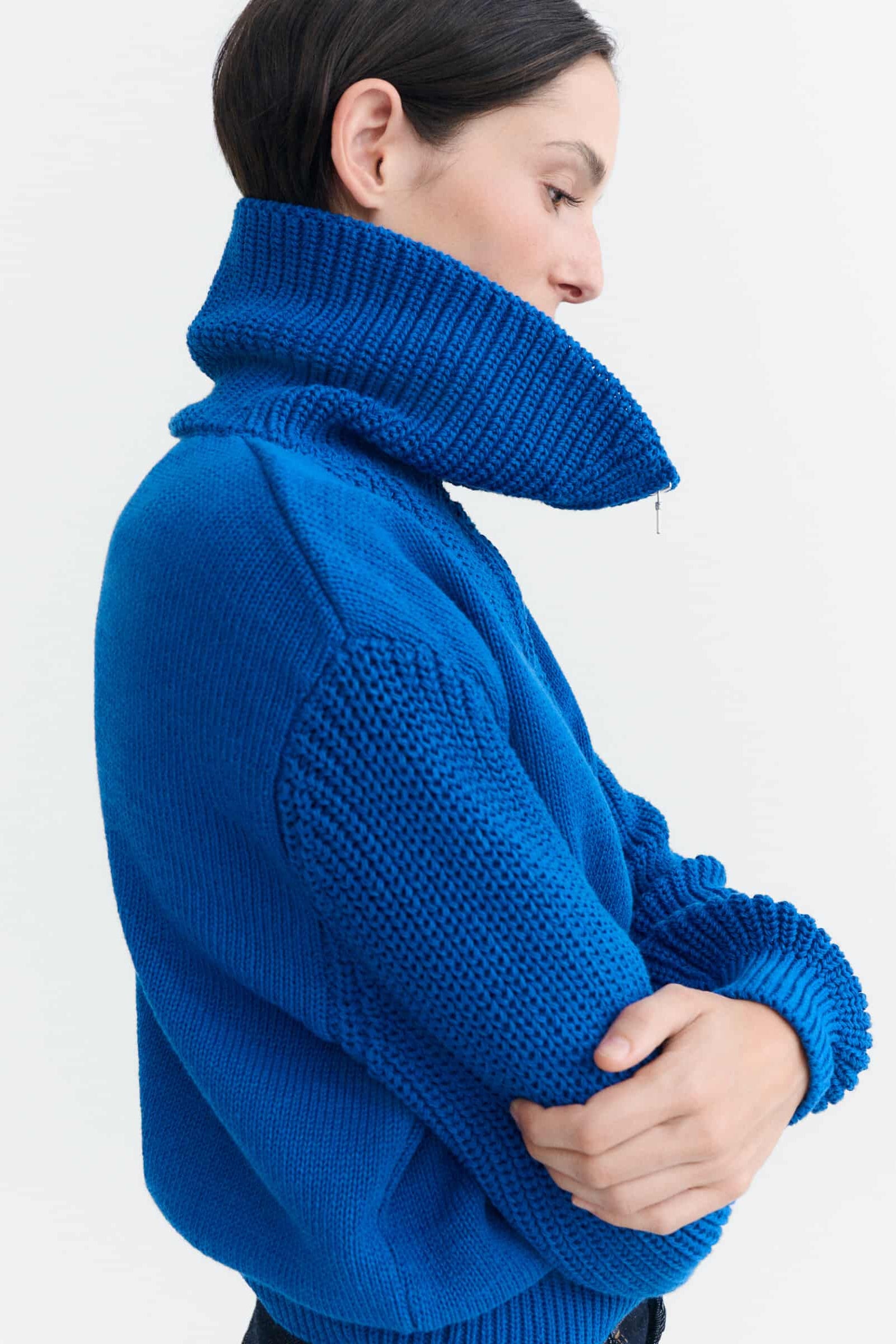 A woman wearing a vibrant royal blue chunky-knit sweater with a high folded collar, shown from a side profile against a plain white background.
