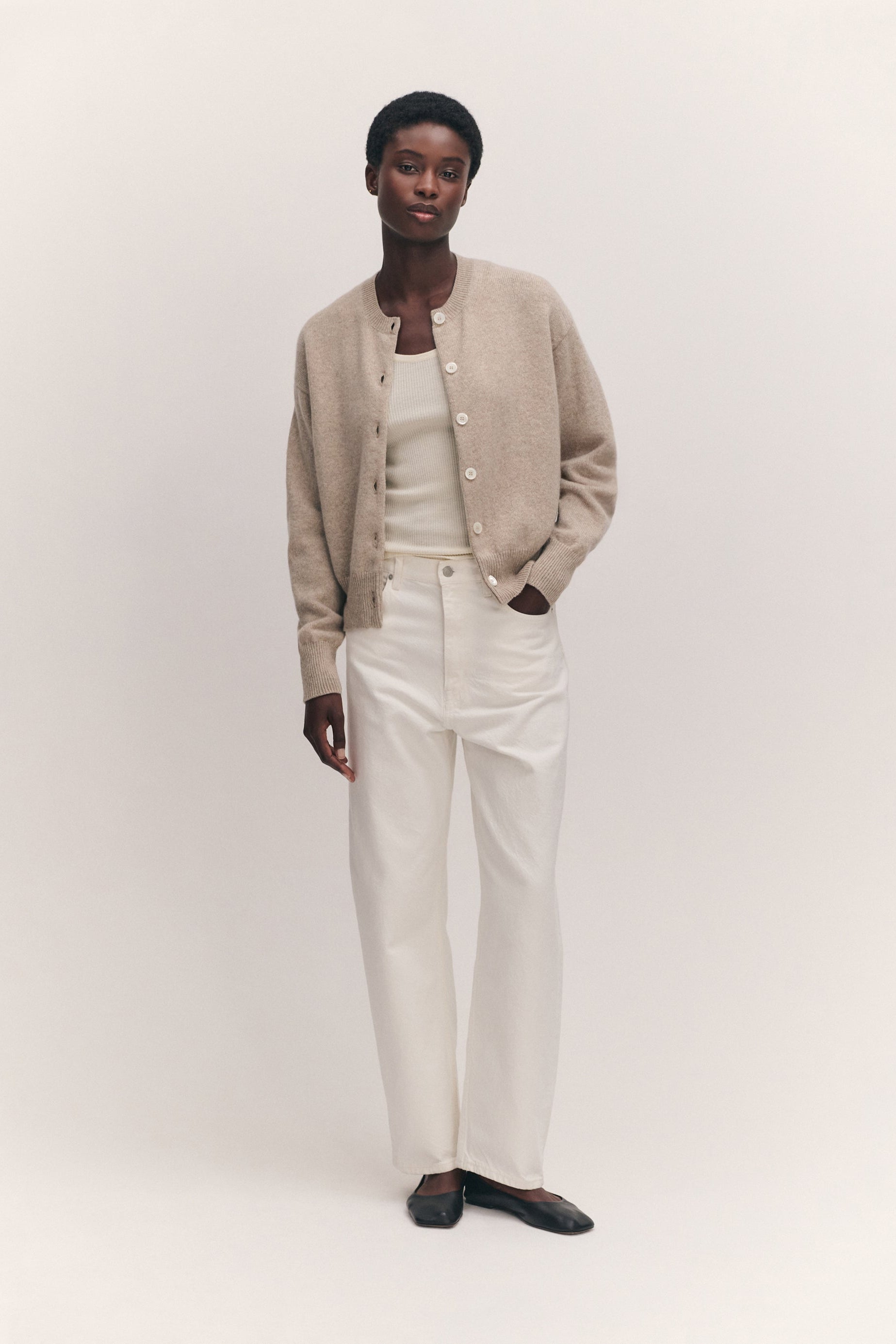 A model stands against a plain light beige studio background wearing a beige cardigan over a white top and white trousers, with black flats.