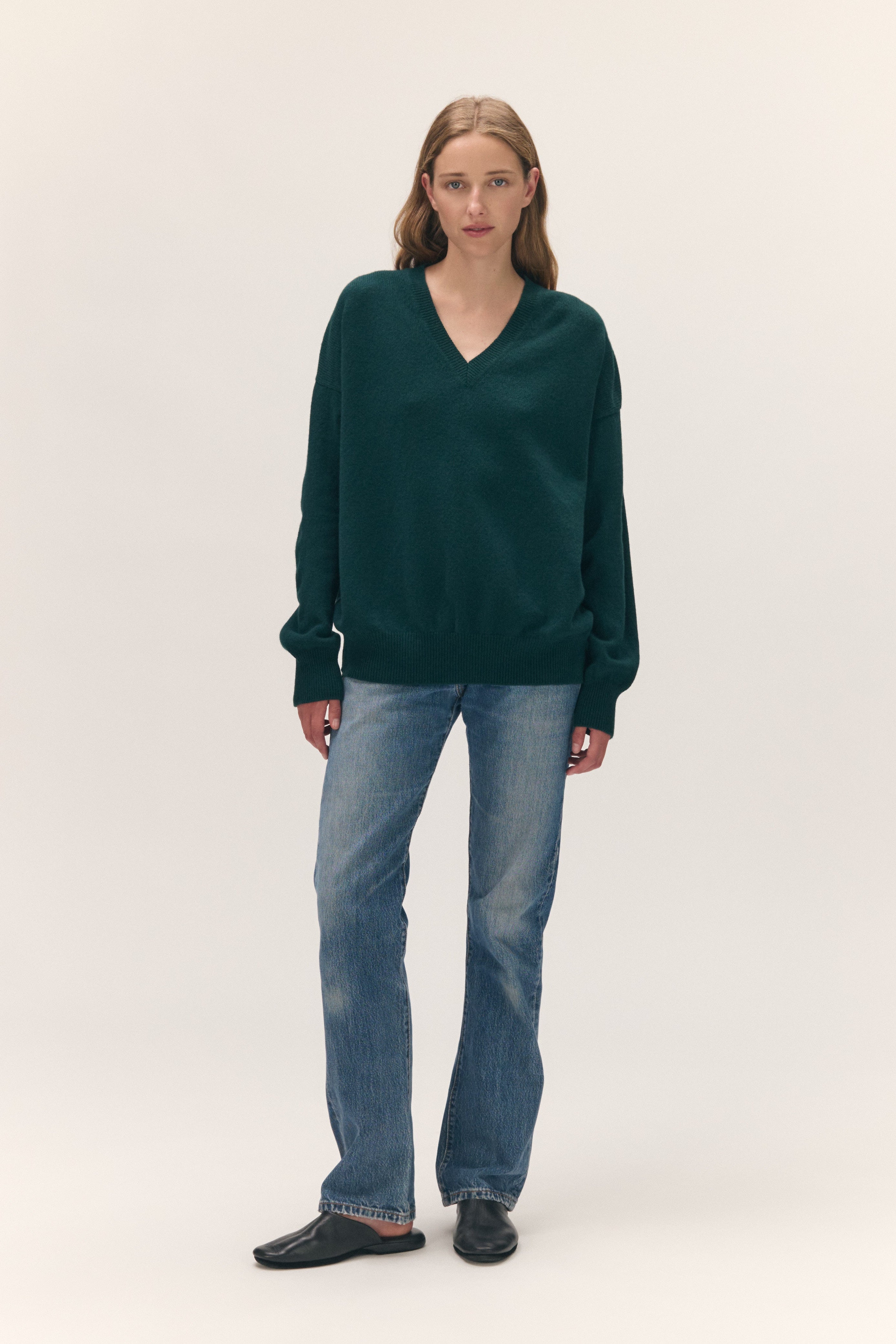 A woman modeling a dark green V-neck knit sweater with blue jeans and black slip-on shoes, standing against a plain light beige studio background.