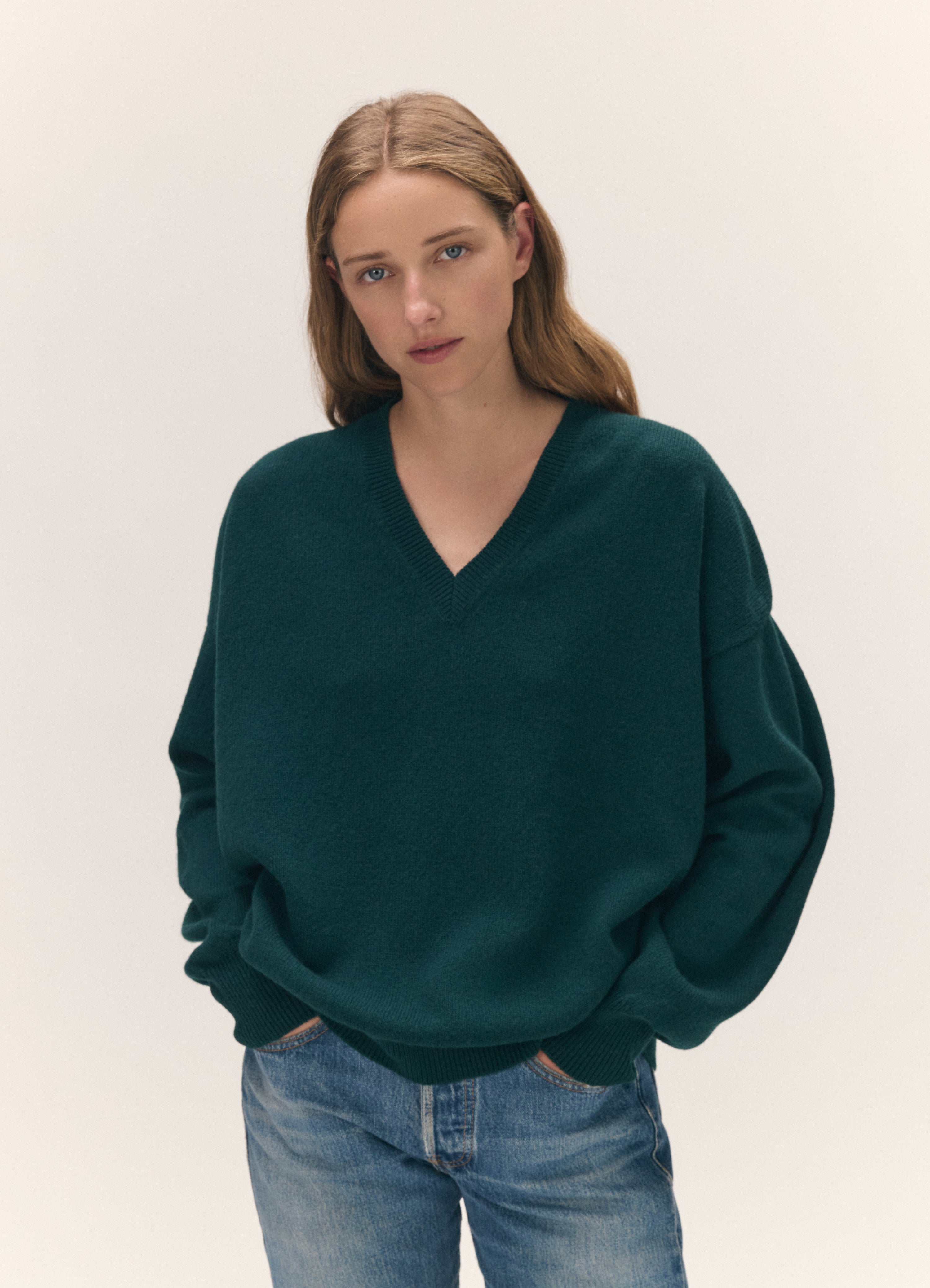 A woman wearing an oversized dark green V-neck sweater and blue jeans, standing against a plain light beige studio background.
