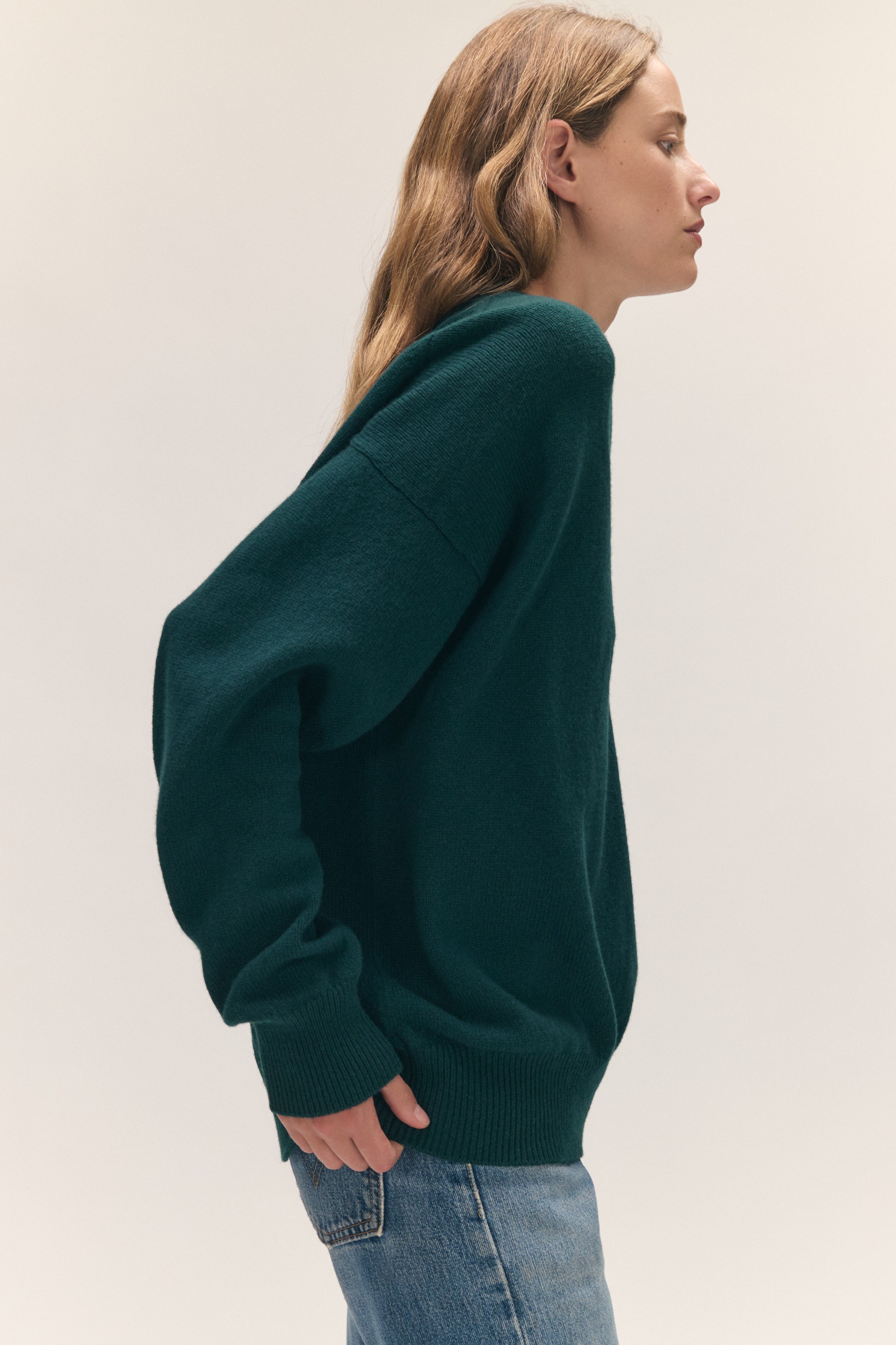A woman shown in side profile wearing an oversized dark green knit sweater and light blue jeans against a plain light gray studio background.