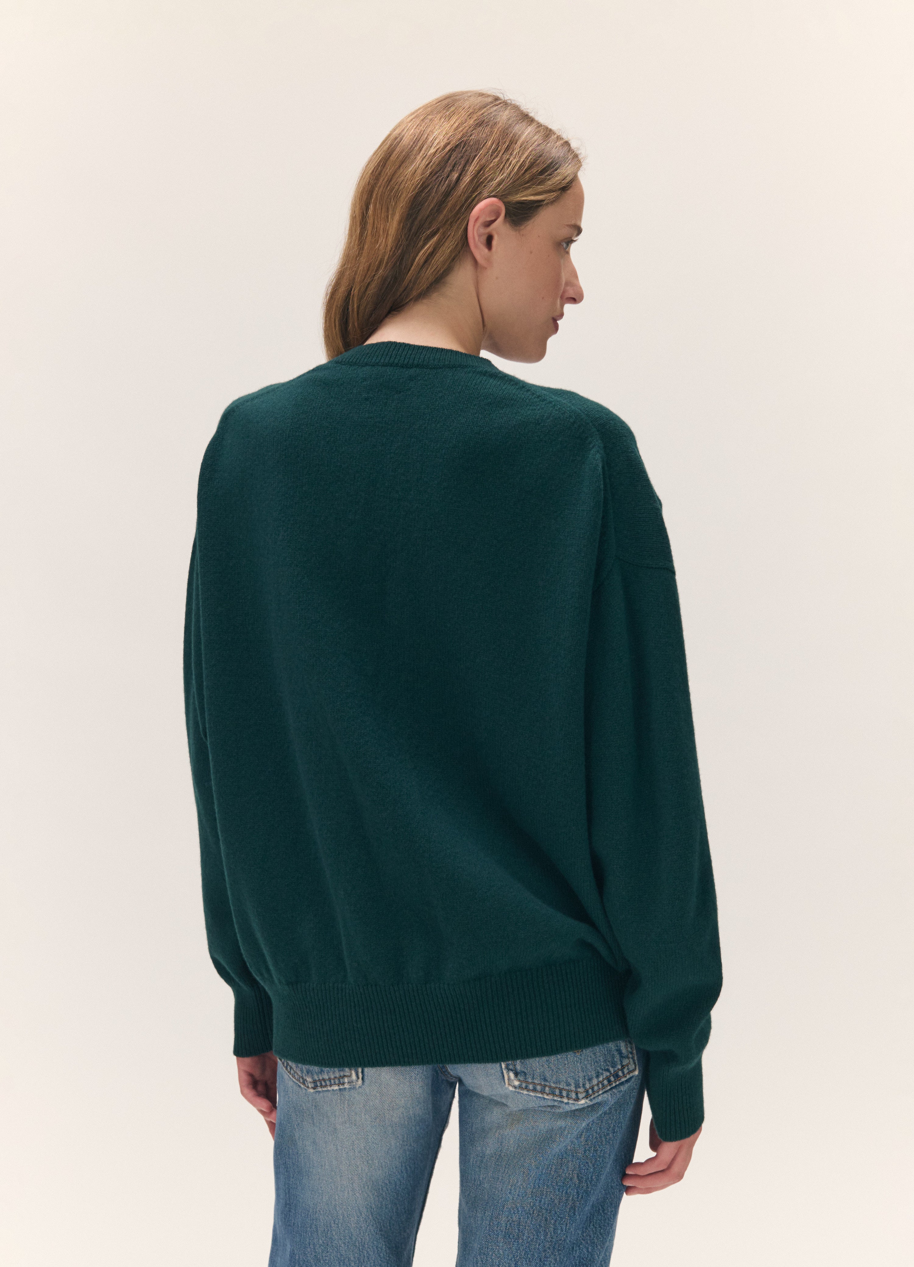 A woman shown from the back wearing an oversized dark green knit sweater and light blue jeans, photographed against a plain off-white studio background.