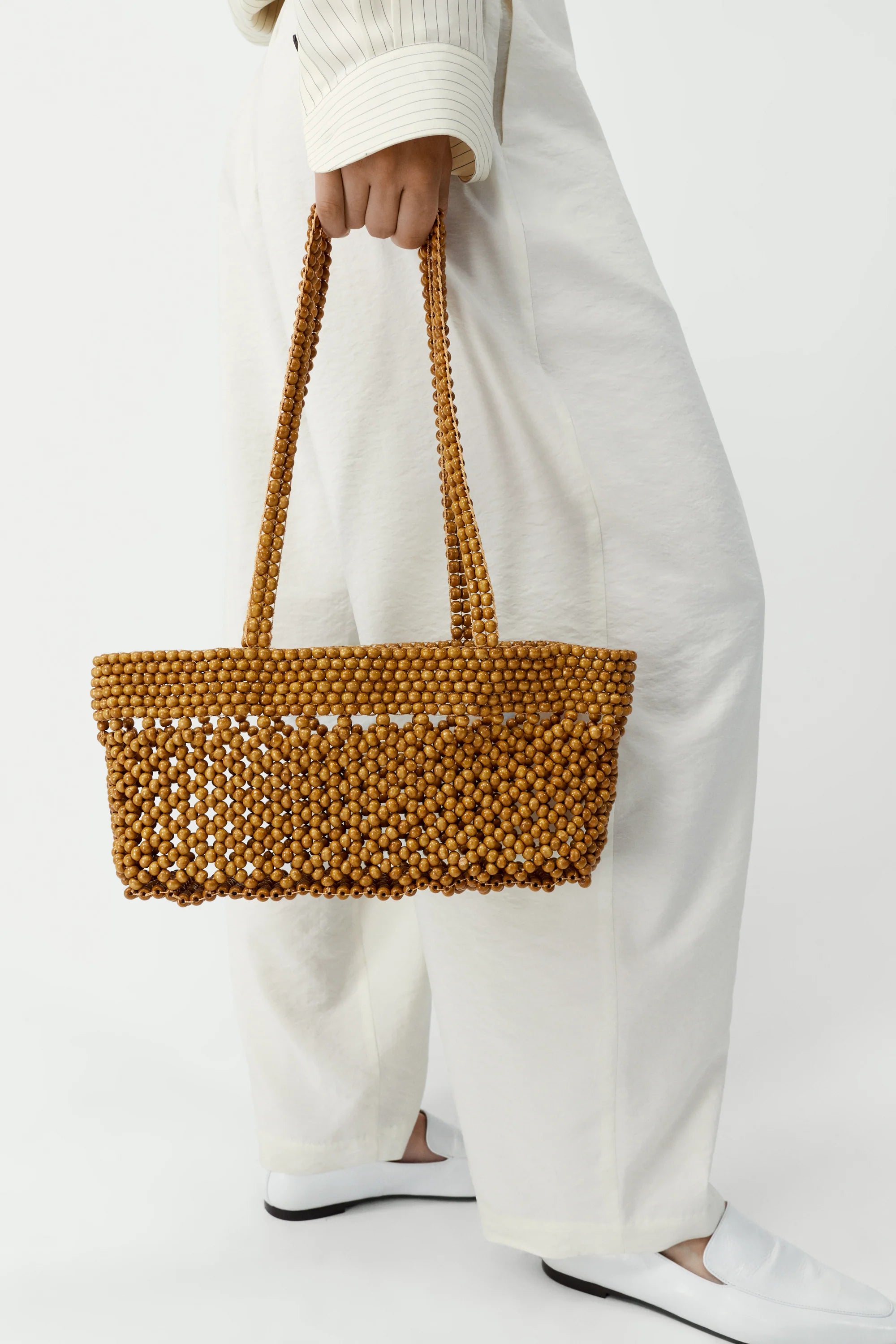 Person holding a beaded brown bag with white pants and a white sweater on a white background
